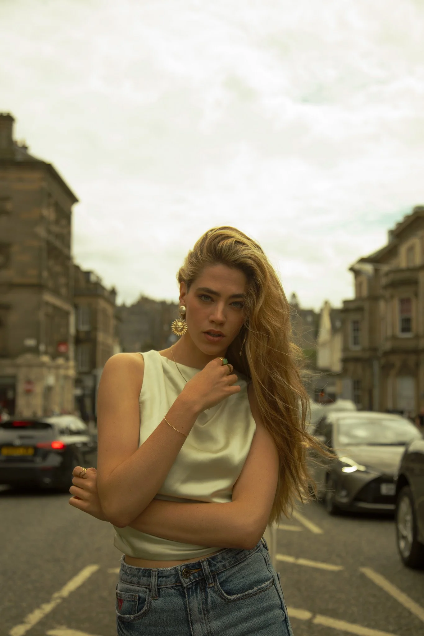 A young woman with long blonde hair and blue eyes standing in the middle of a city street, wearing a sleeveless satin top and high-waisted jeans, with buildings and cars in the background on a cloudy day.