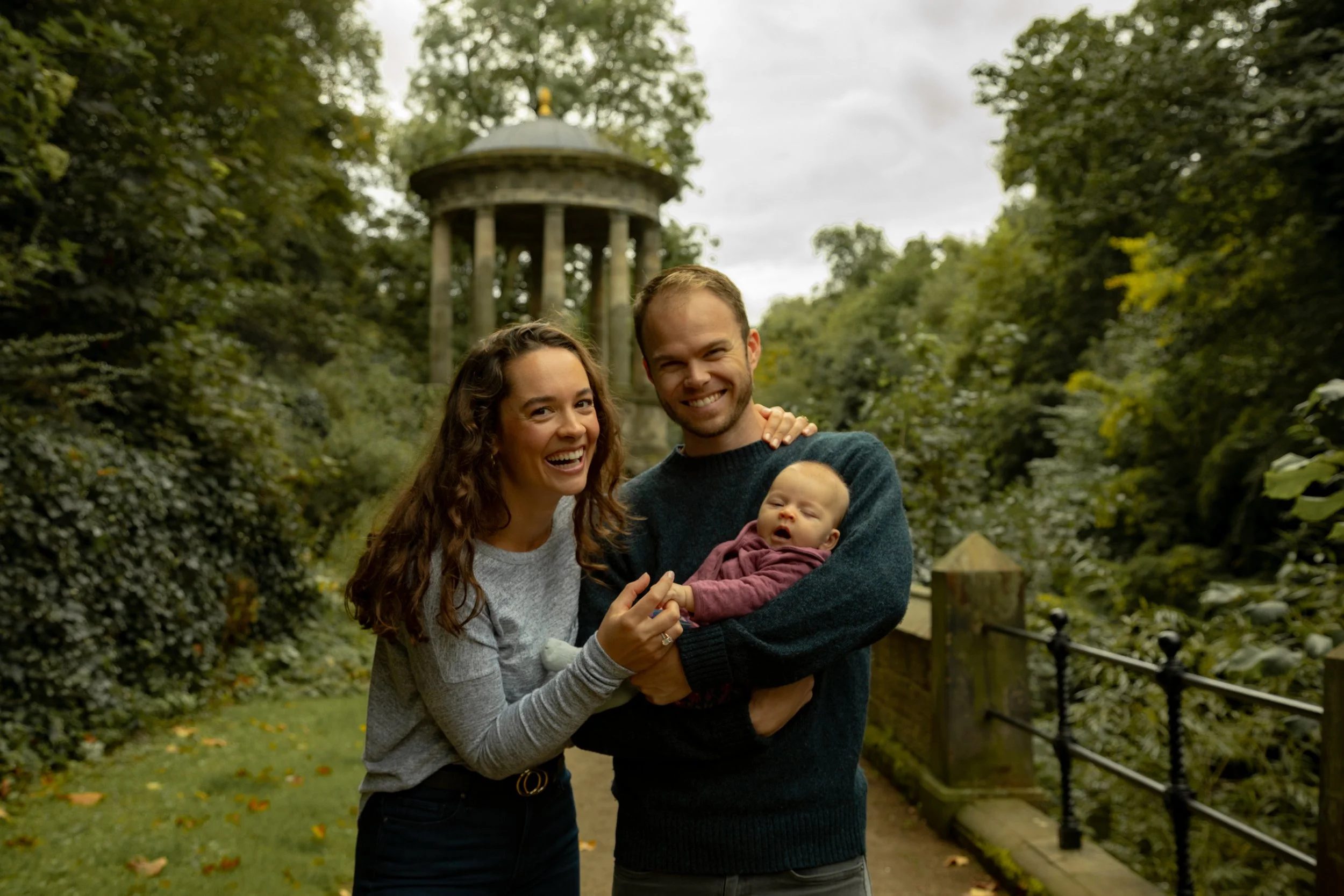 A happy family of three, a woman, a man, and a baby, standing outdoors in a park with lush greenery and a gazebo in the background, smiling at the camera.