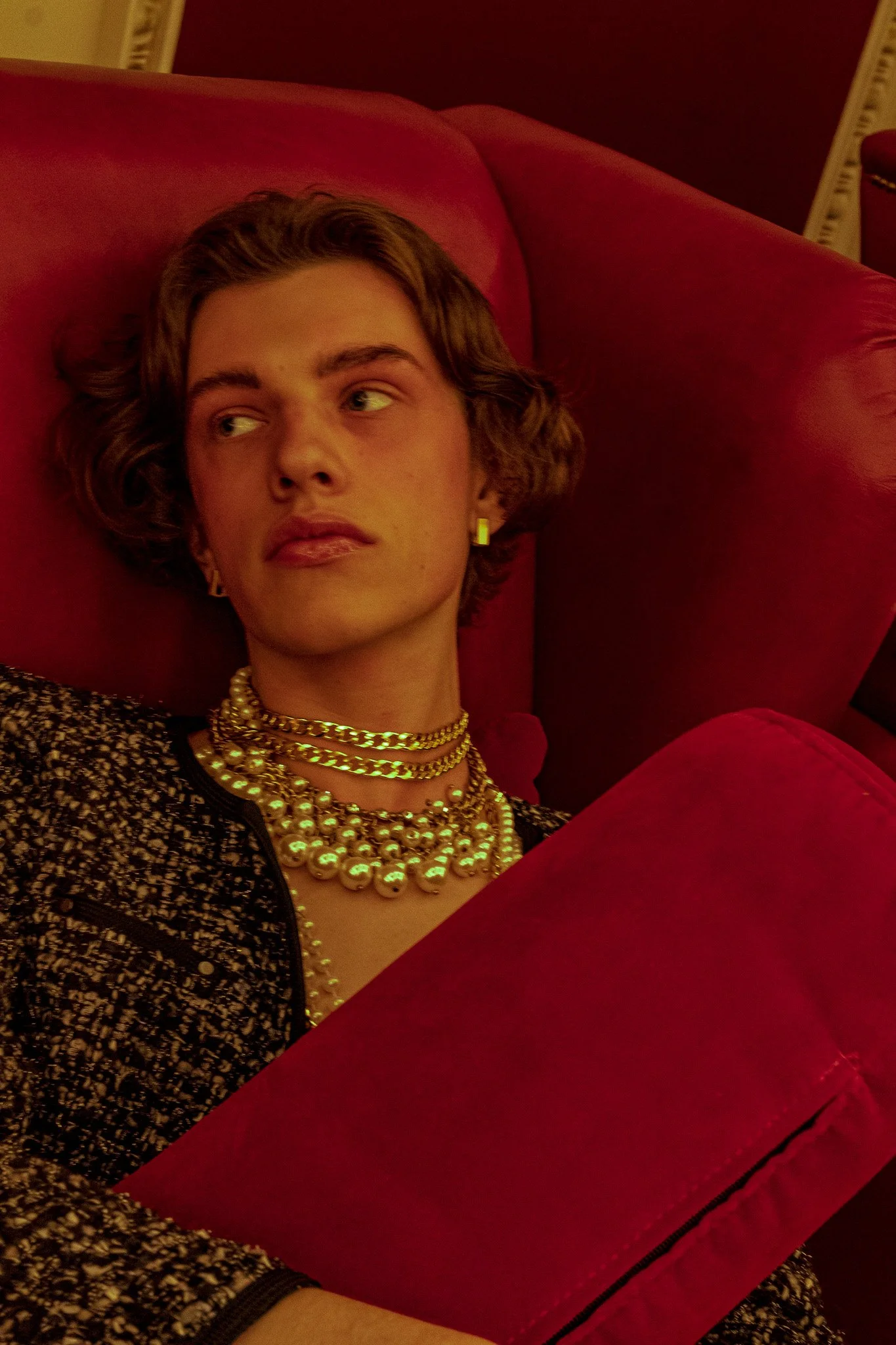 A woman with short, curly hair lying on a red couch, wearing multiple pearl necklaces, gold chains, gold earrings, and a patterned blazer.