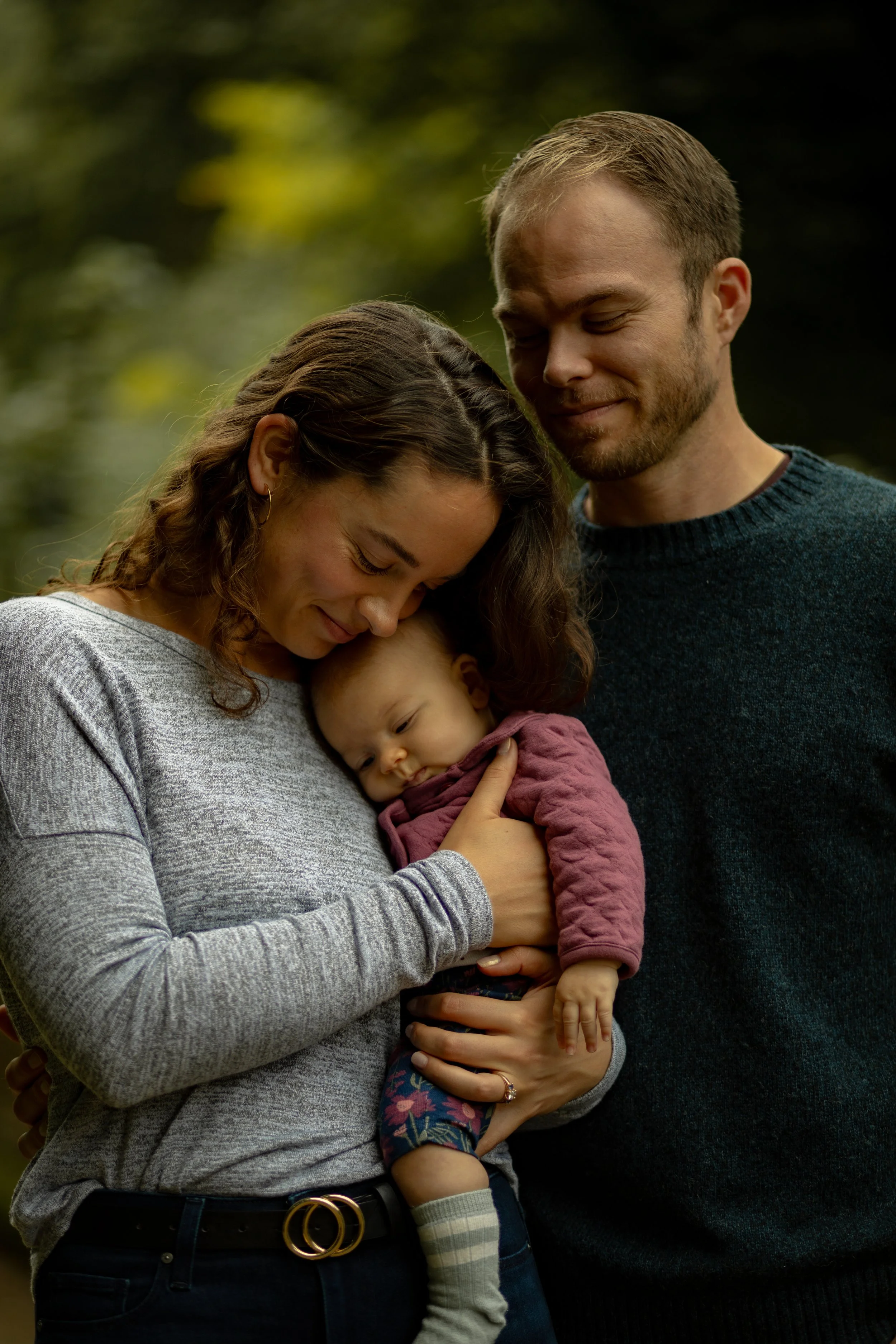 A family of three, a woman, a man, and a baby, sharing a tender moment outdoors. The woman is holding the baby close to her chest, and both are smiling softly with eyes closed. The man is leaning his head gently on the woman's, also with a loving expression.