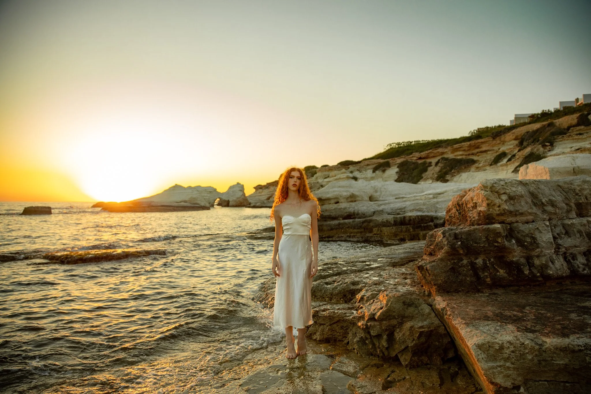 A woman with red curly hair in a white dress standing on the rocky shoreline during sunset.