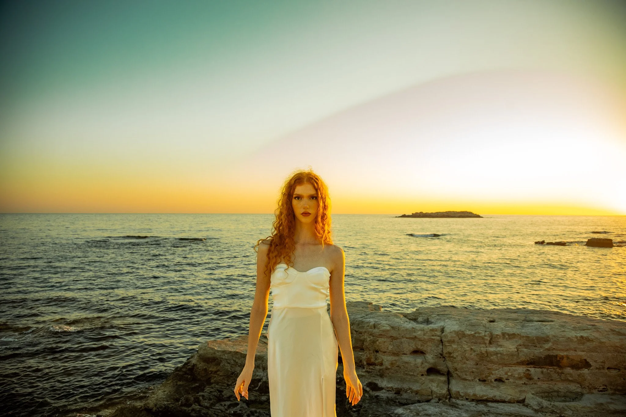 A woman with long curly red hair in a white dress standing on the rocks at the beach during sunset.
