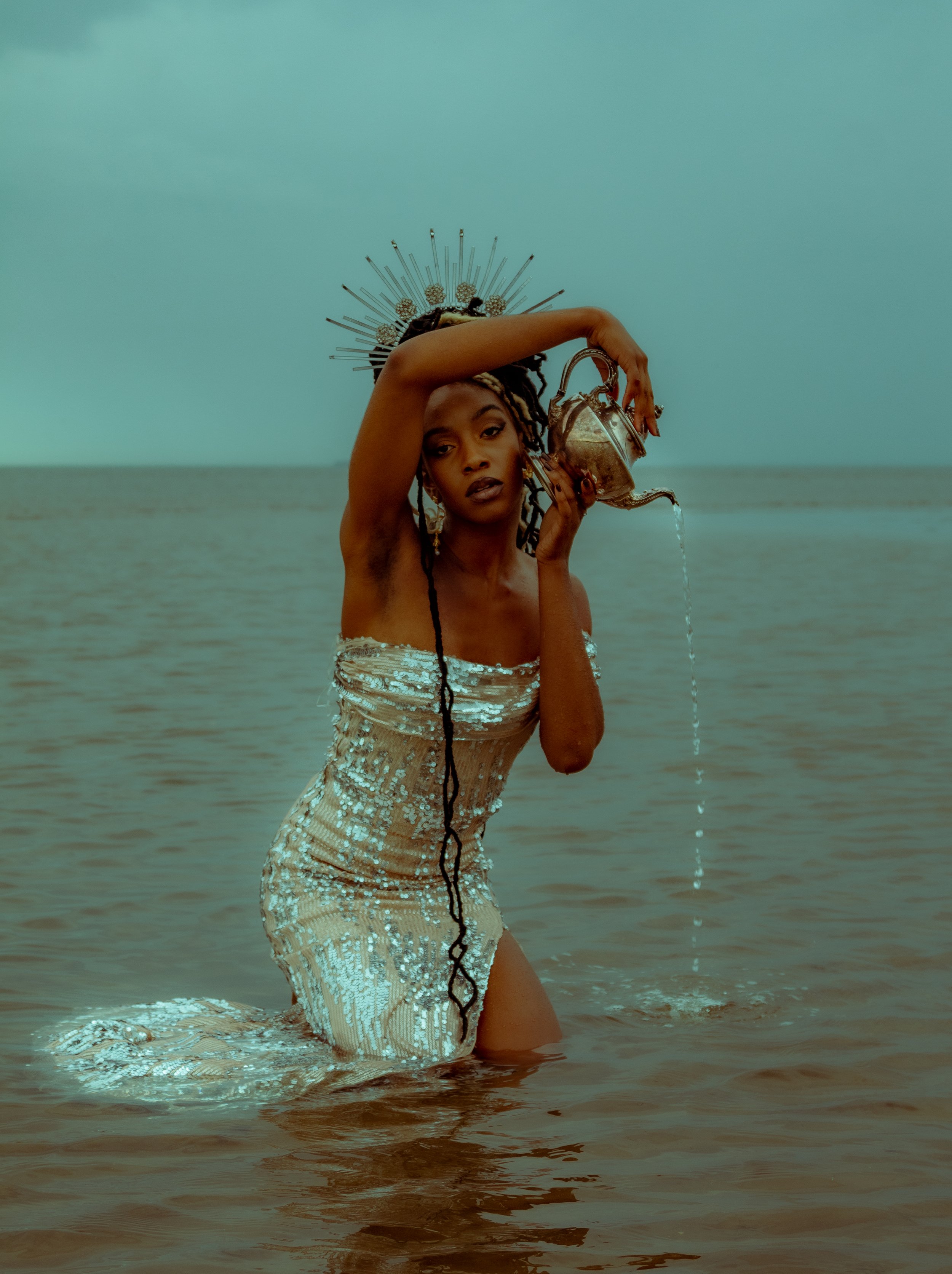 A woman standing knee-deep in water, wearing a strapless sequined dress and a decorative headpiece with spikes, holding a metallic teapot from which water is pouring.