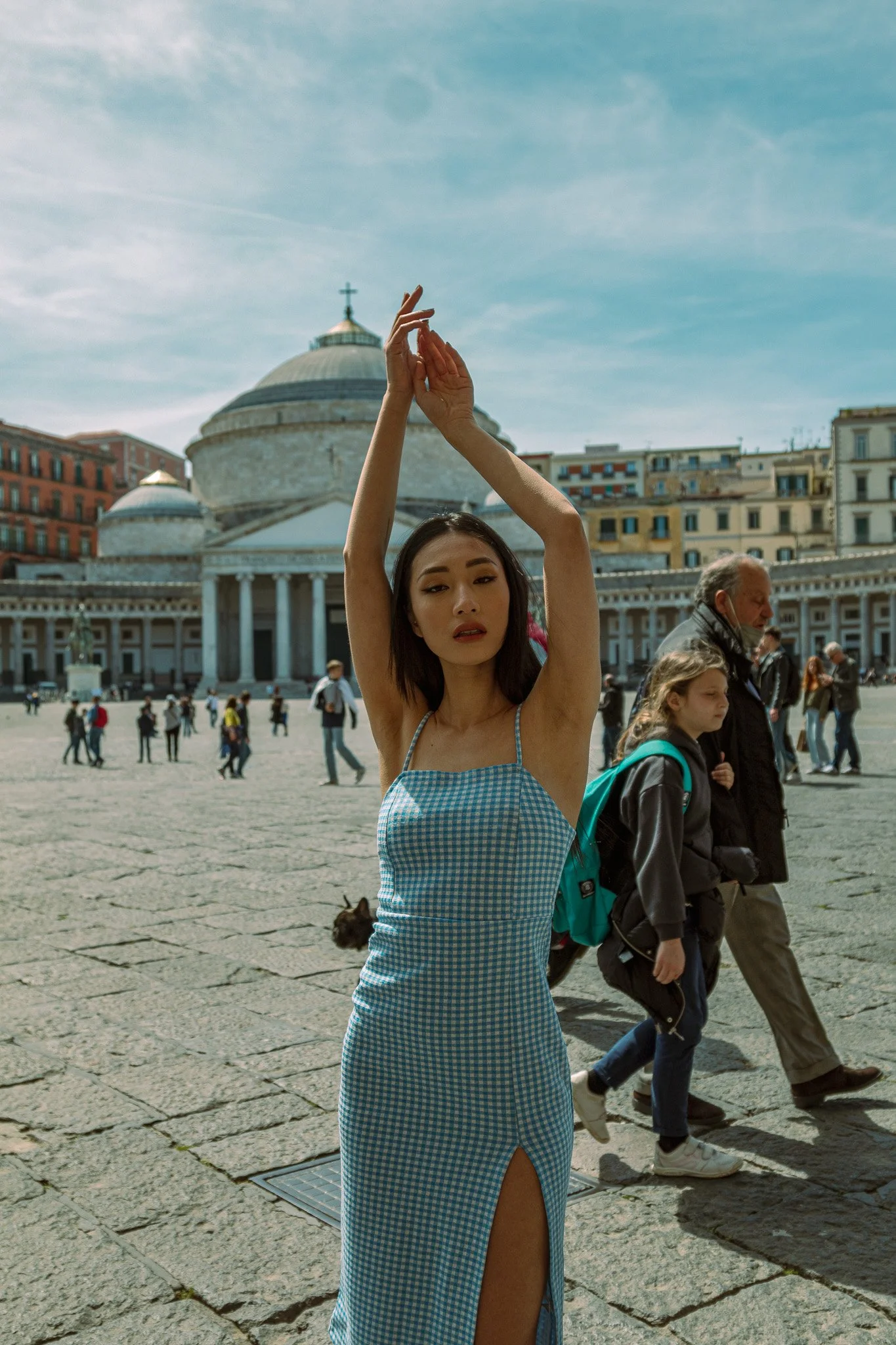 A woman with long dark hair and a checkered blue dress standing with arms raised in front of a historic building with a large dome, with other people walking in the background.
