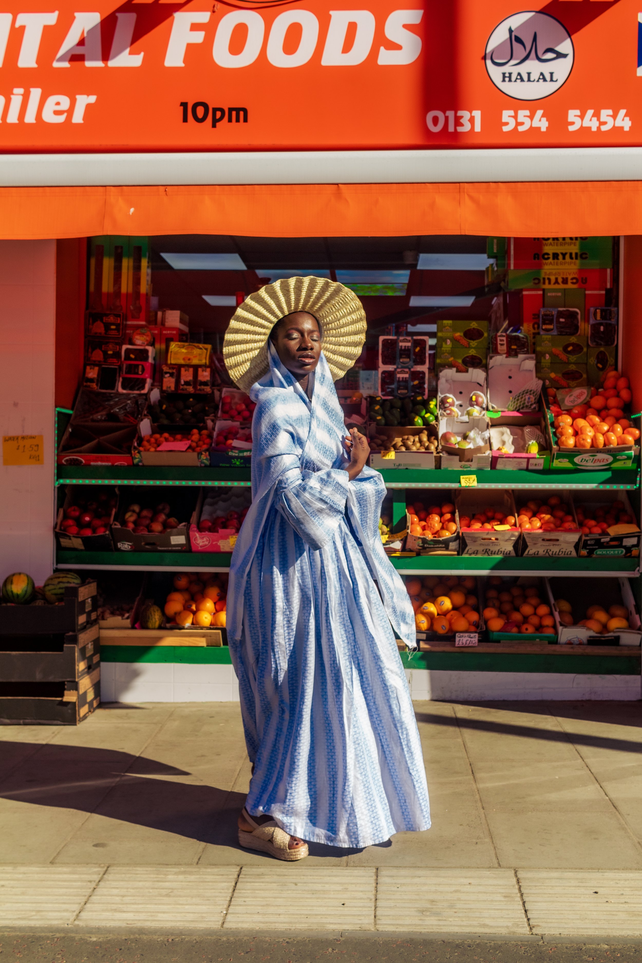 A woman dressed in traditional attire and wearing a large straw hat, standing in front of a fruit stand at a grocery store.