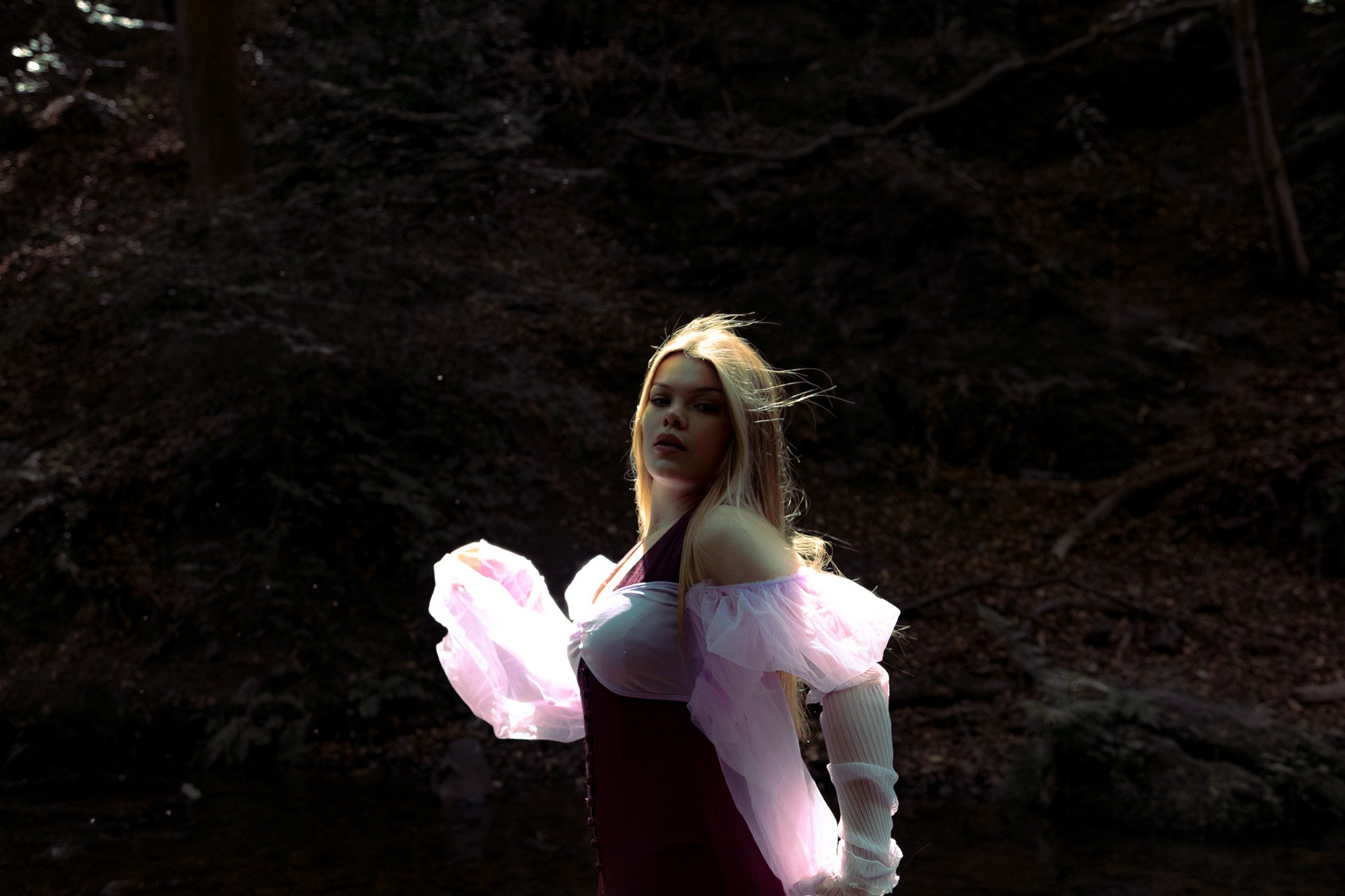 A woman with long blonde hair, dressed in a dark outfit with pink ruffled sleeves, stands in a dark outdoor setting, illuminated by light that highlights her face and upper body.