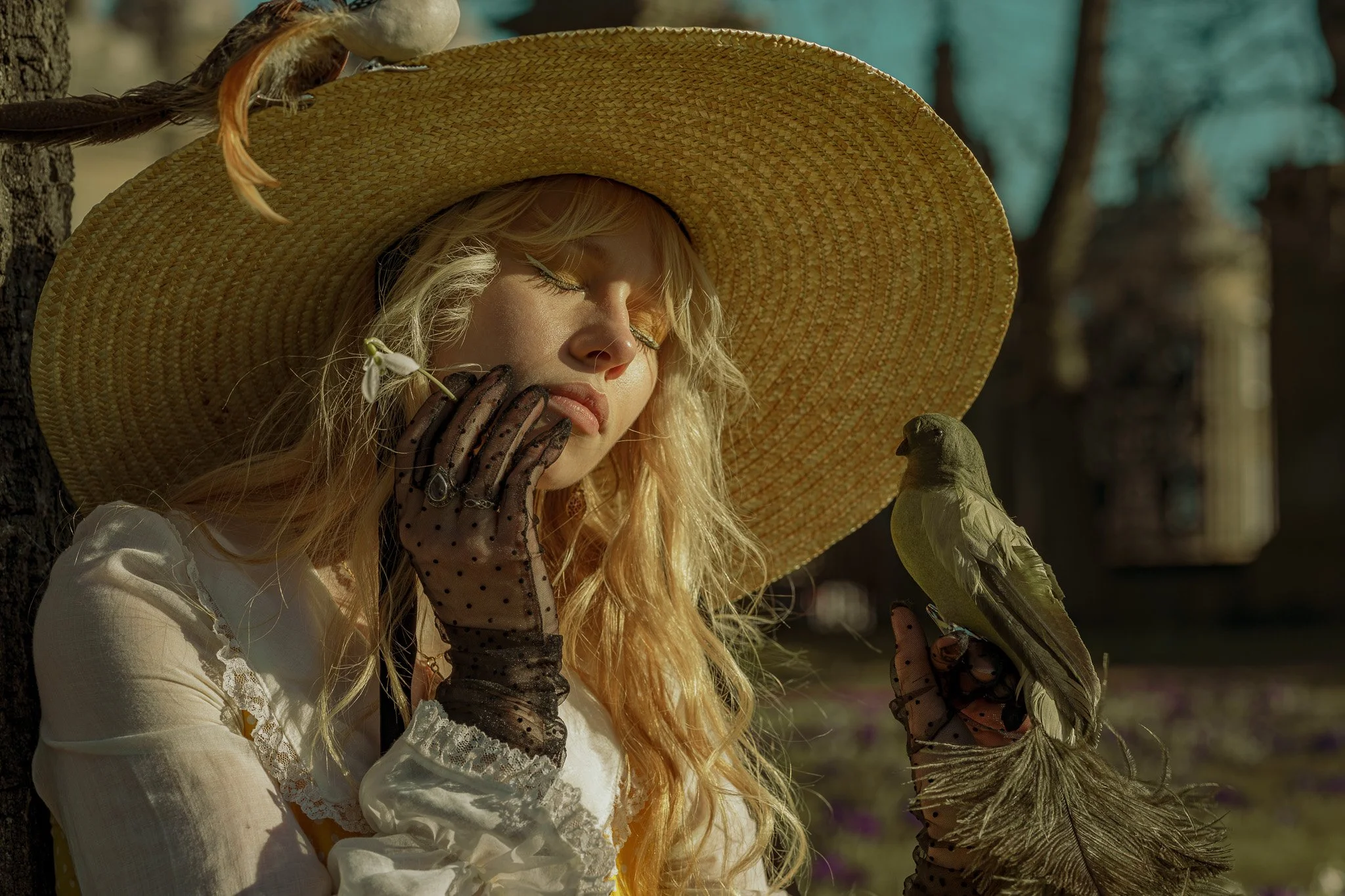 A young woman with long blonde hair wearing a large straw hat, lace gloves, and a vintage white dress, holds a bird on her glove while her eyes are closed and her face is turned slightly downward, outdoors in a natural setting.