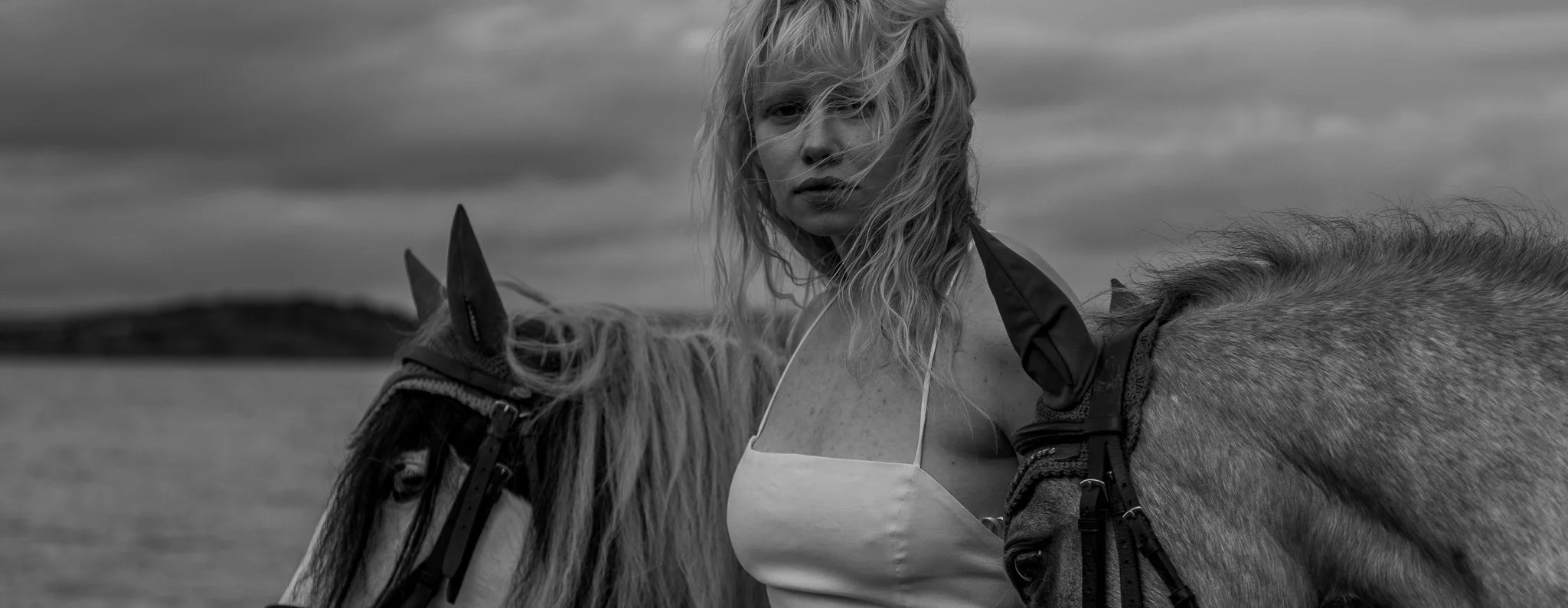 Black and white photo of a woman with wavy hair standing next to a horse beside a body of water, with clouds in the sky.