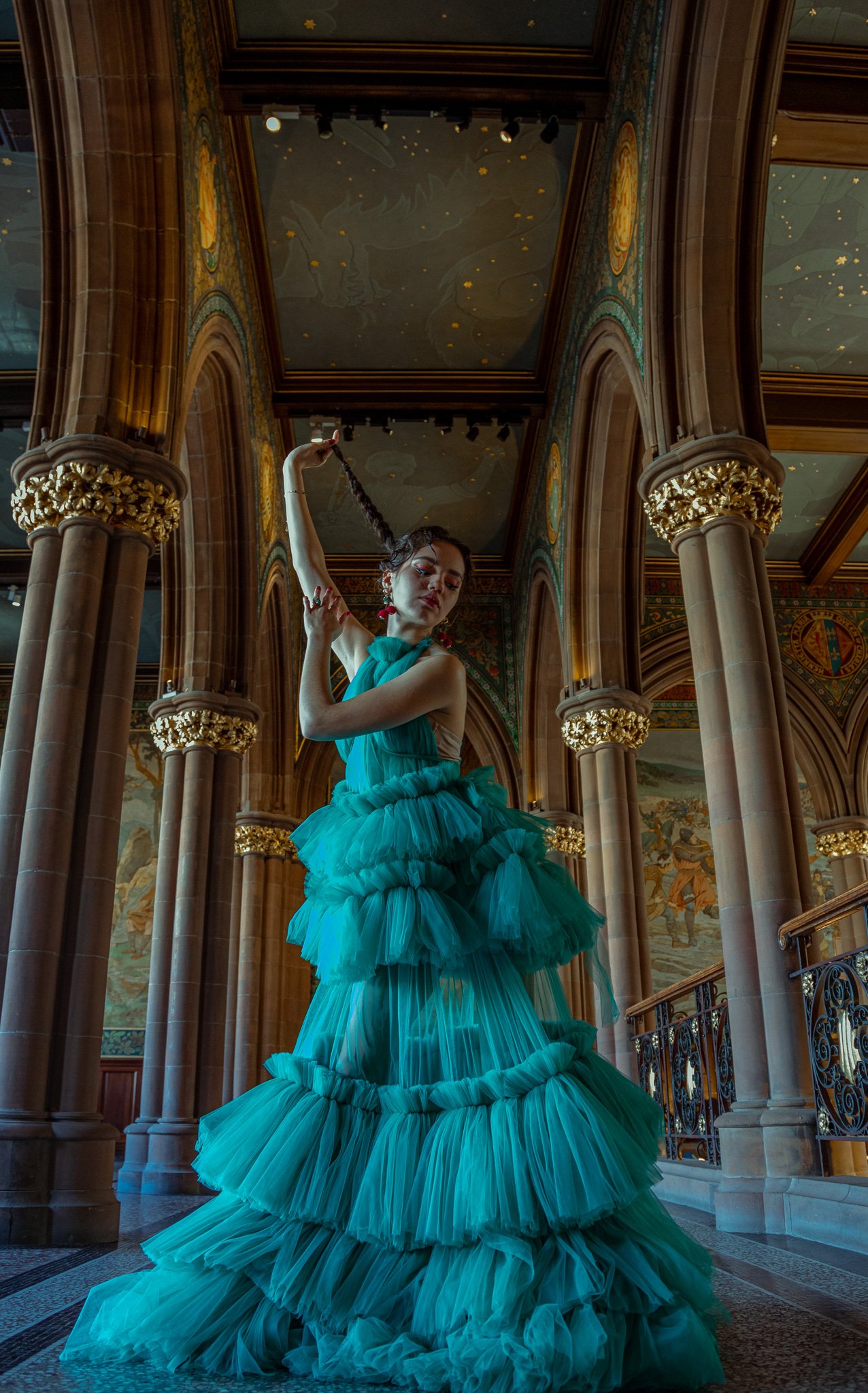 A woman in a turquoise, ruffled, layered gown with a high neckline, standing in an ornate, historic building with tall columns and mural paintings on the walls. She has long dark hair styled in a braid, which she holds in one hand, and her eyes are c