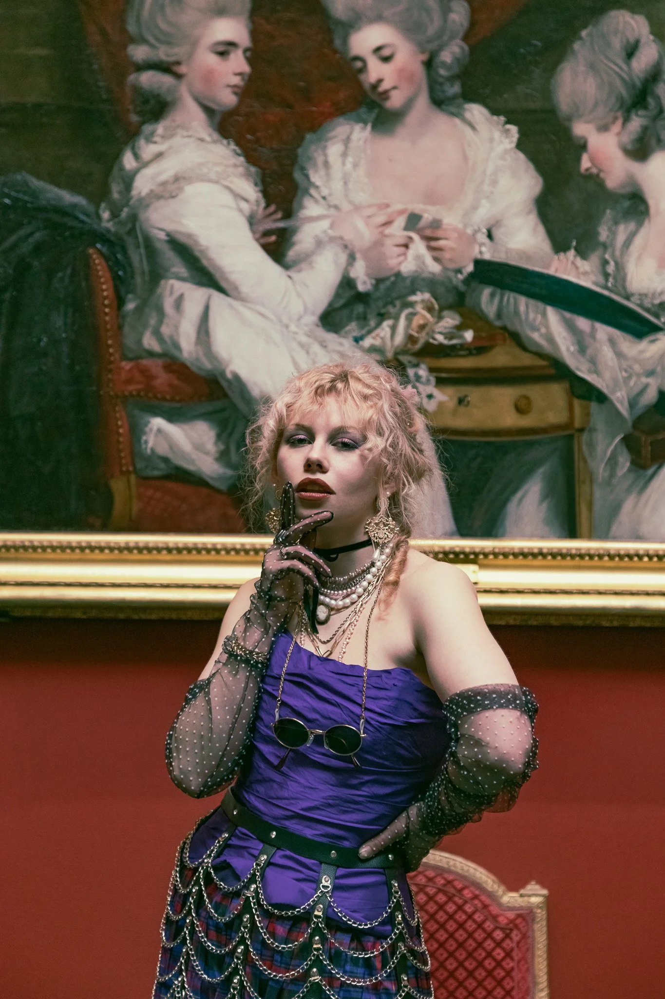 A woman with curly blonde hair dressed in punk fashion, wearing jewelry, a purple top, and plaid pants, standing in front of a large classical painting of women in historical attire.