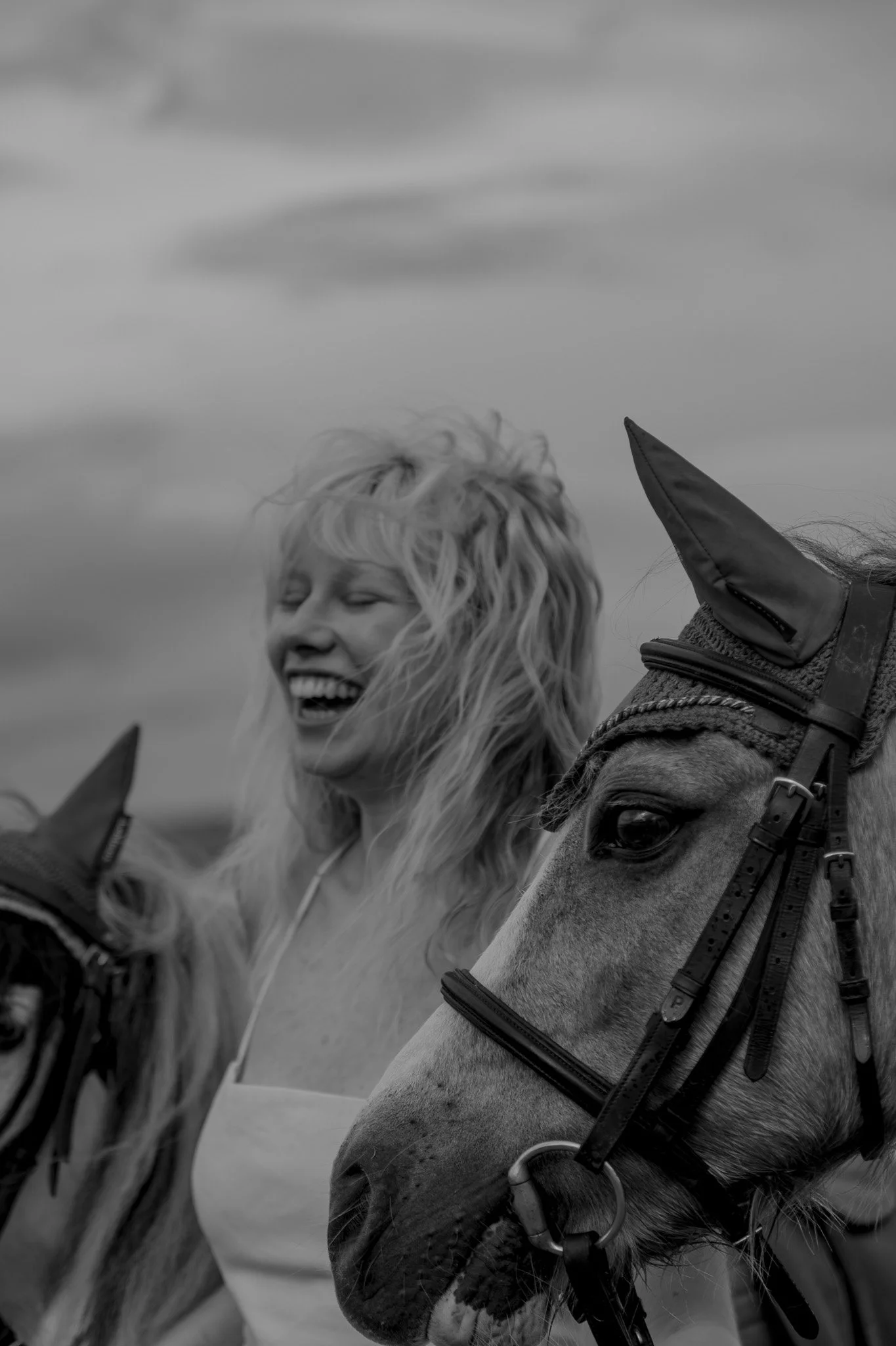 A woman with curly blonde hair laughing with two horses, one on each side, wearing bridles and ear covers, outdoors with a cloudy sky in the background.