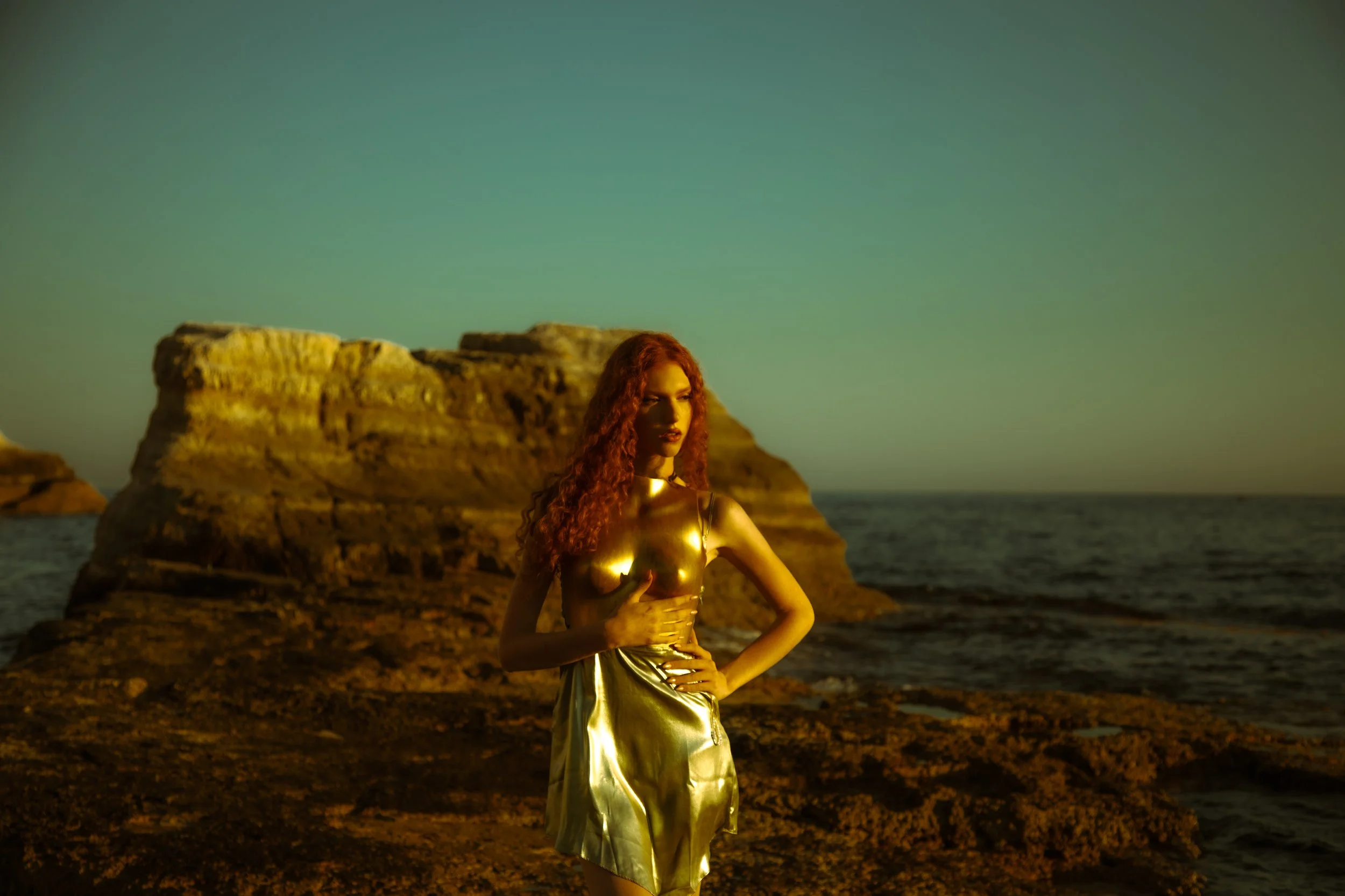 A woman with long, curly red hair stands on a rocky beach at sunset, wearing a shiny gold dress.
