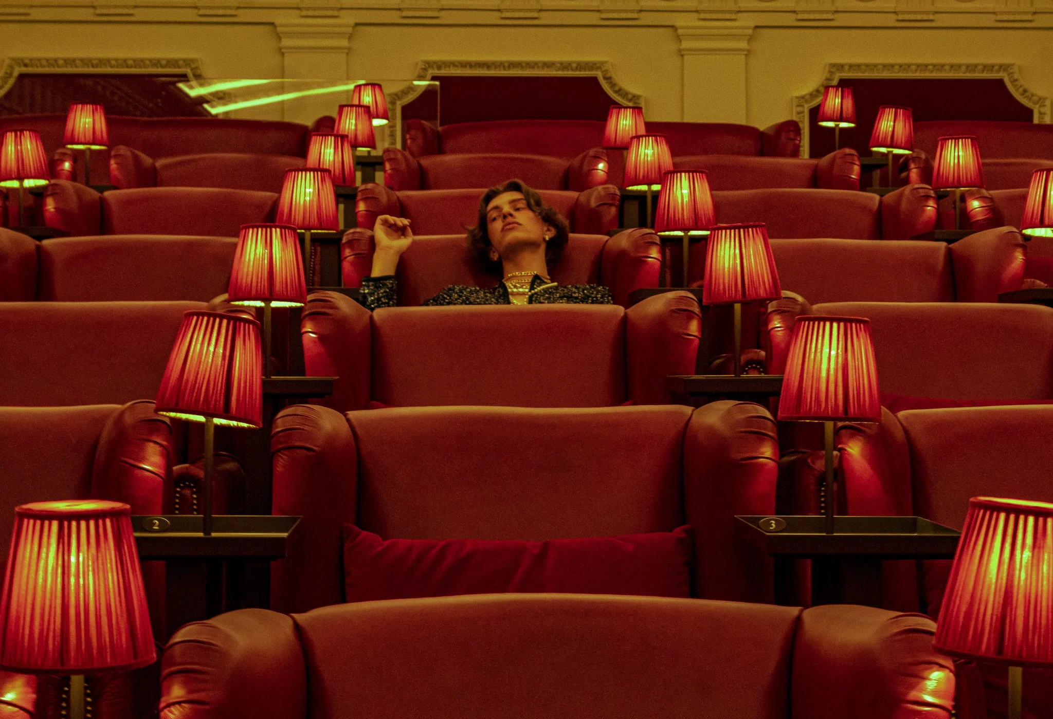 A woman relaxing alone in an empty theater or cinema, surrounded by red armchairs and red lamps, with a dark ornate wall in the background.