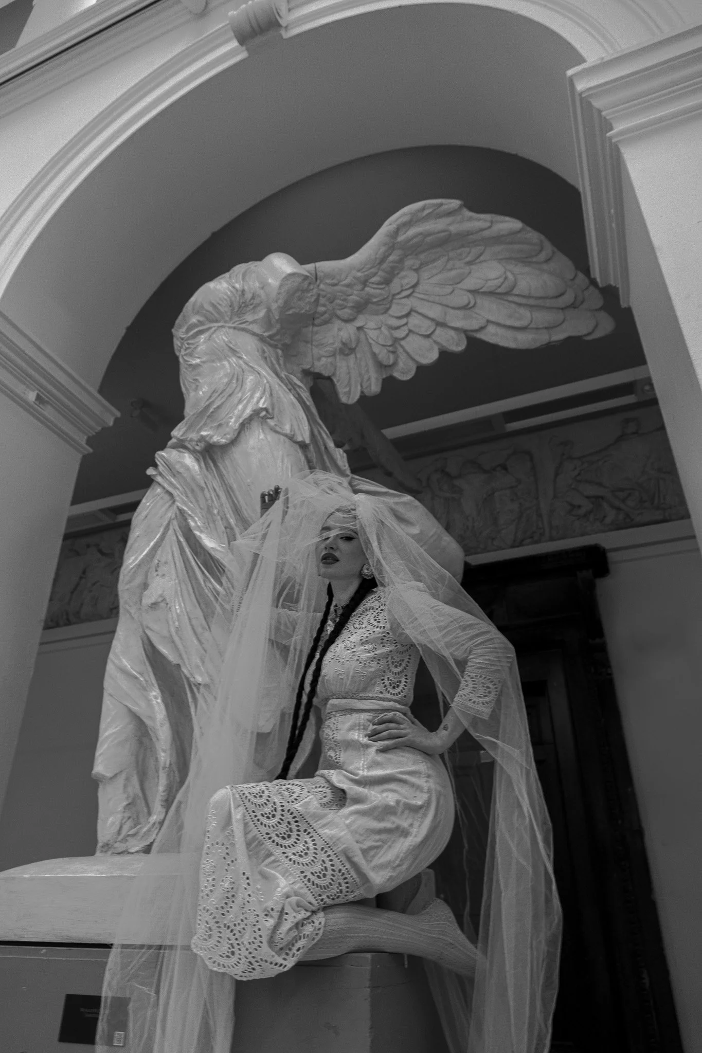 A woman dressed in a lace dress with a veil kneeling in front of a large angel sculpture with wings in a museum or gallery setting.
