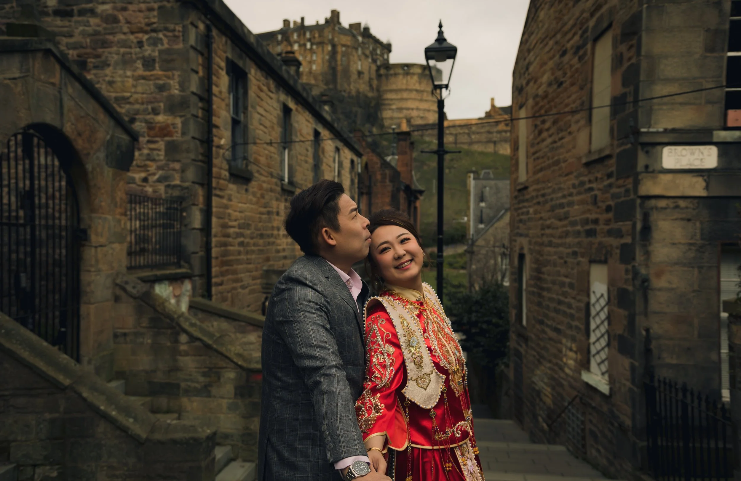 A couple standing on a narrow street with historic brick buildings and a castle on a hill in the background, A man in a gray plaid suit and a woman in traditional red and gold attire smiling and holding hands.