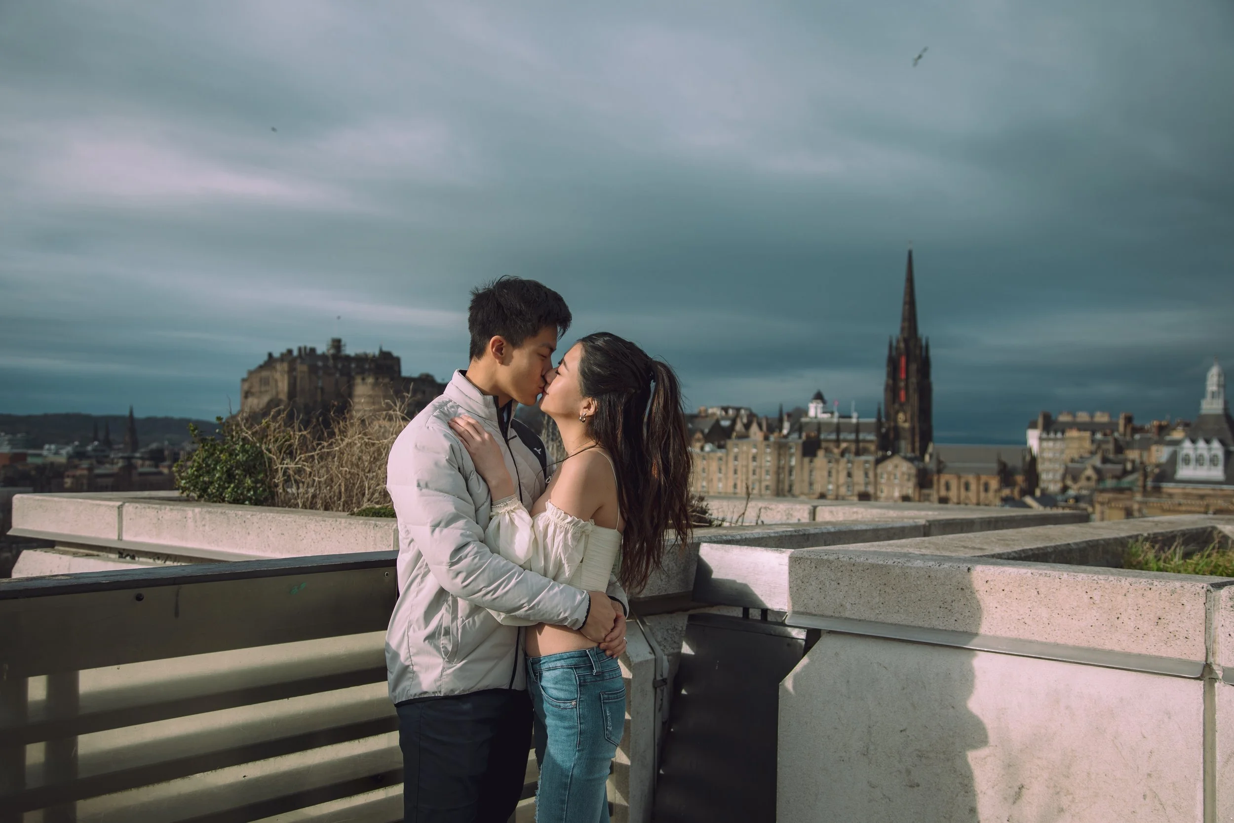 A young couple sharing a kiss on a city rooftop with historic buildings and a castle in the background.