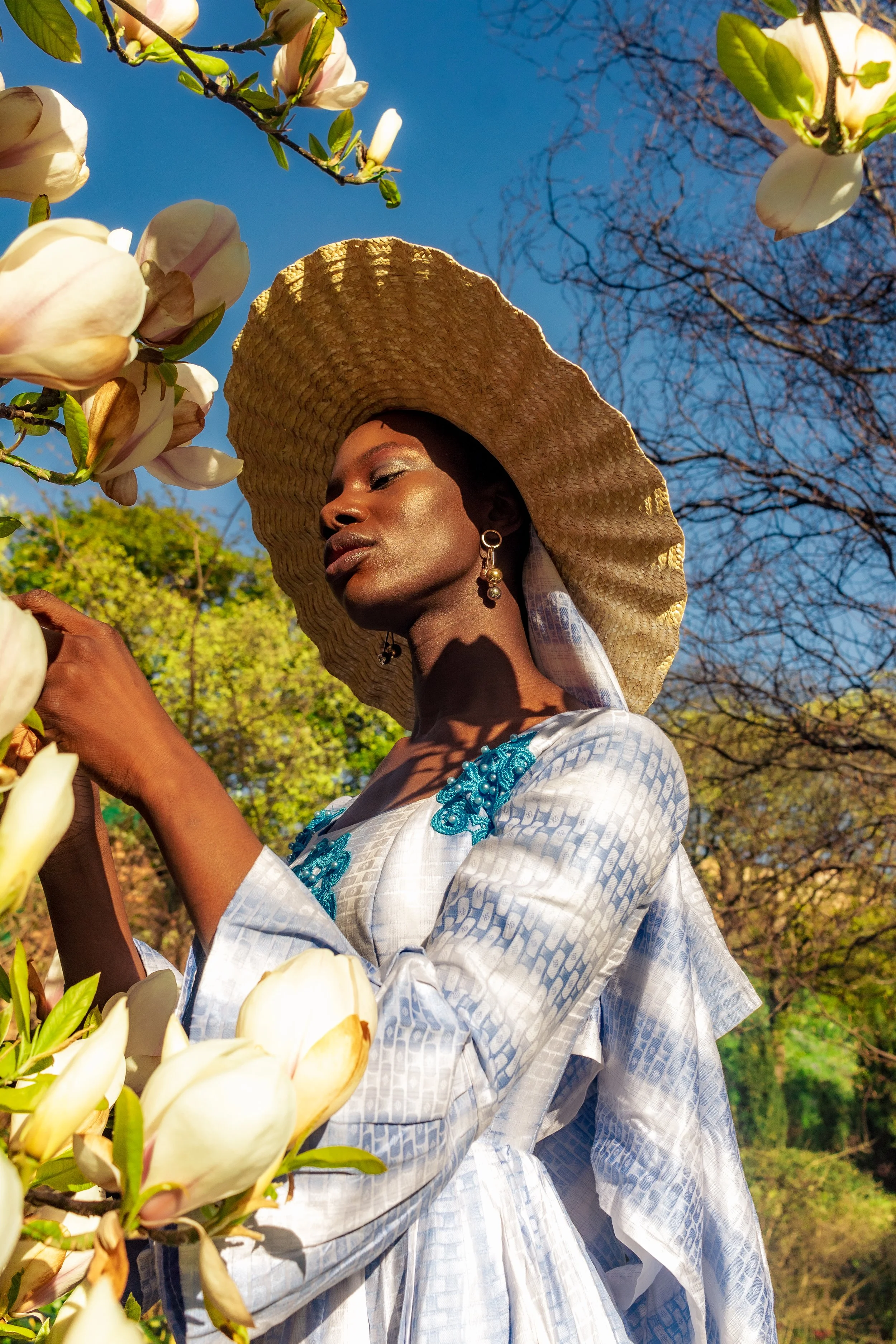 A woman with dark skin wearing a large straw hat, white dress with blue embroidery, and earrings, stands outdoors among white magnolia flowers, with trees and a blue sky in the background.
