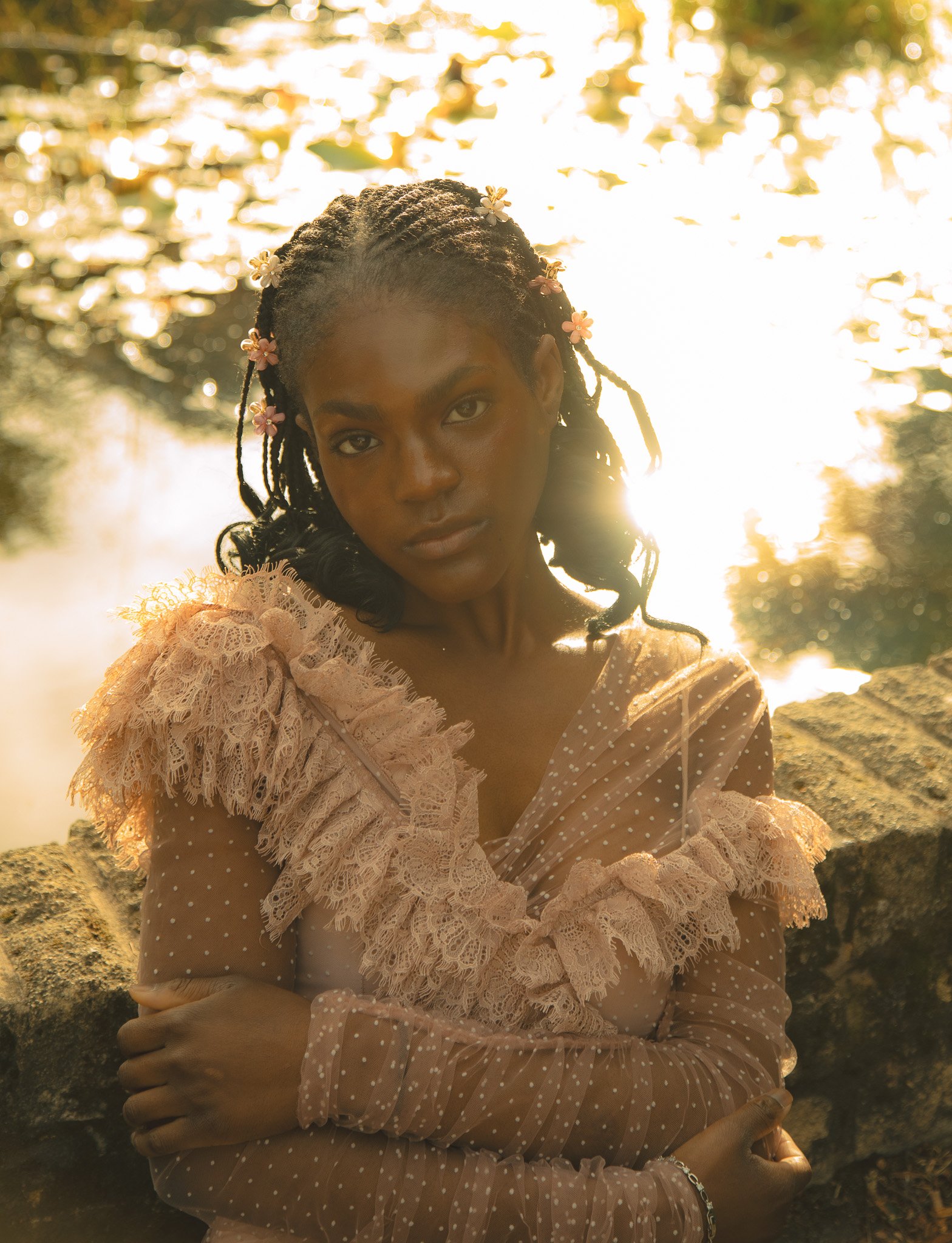 A woman with braided hair decorated with small flowers, wearing a sheer, polka-dotted, ruffled dress, standing outdoors near a stone wall, with sunlight glowing behind her and water in the background.