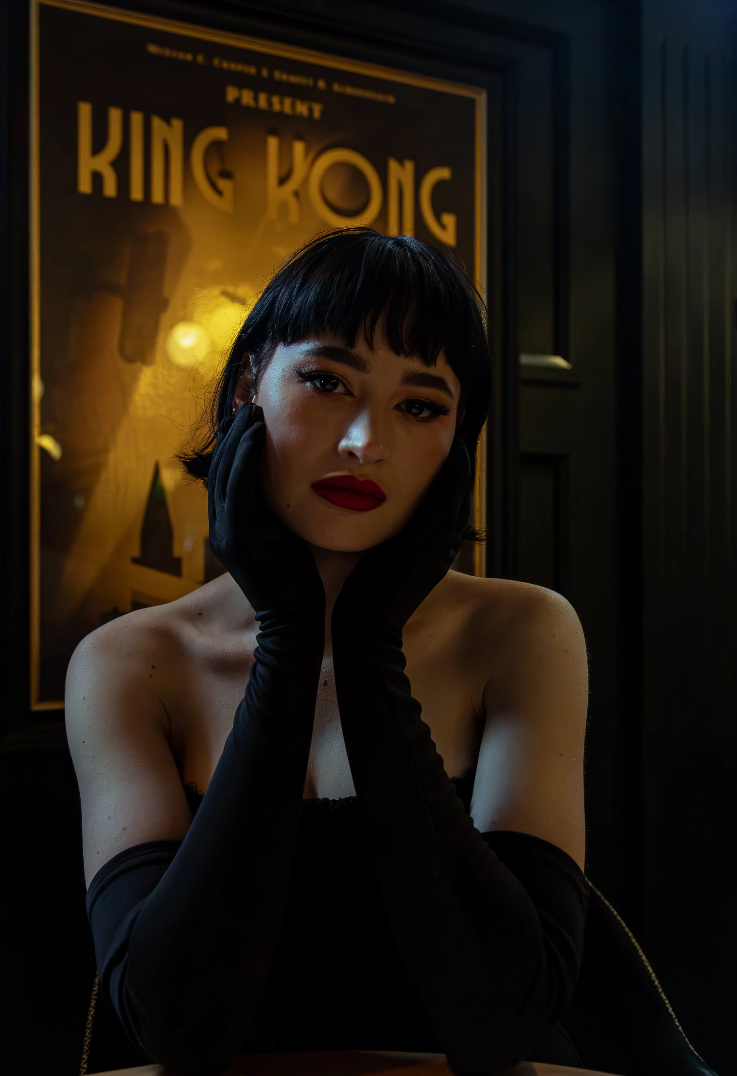 A woman with short black hair, red lipstick, and dark eye makeup posing with her hands on her cheeks, wearing black gloves. She is sitting in front of a vintage King Kong movie poster in a dark, stylish room.