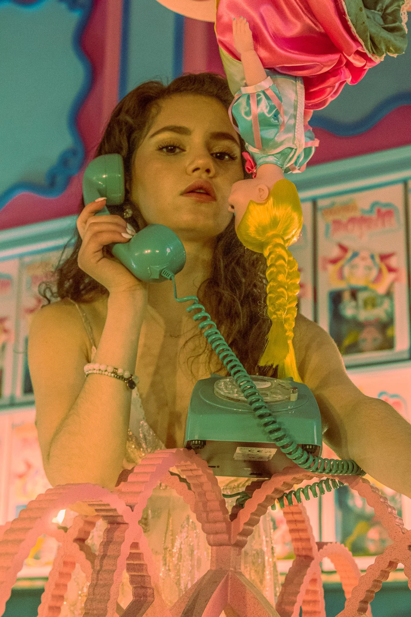 A woman with curly hair talking on a teal rotary phone, holding a blonde doll with a braid, surrounded by colorful decor in pink and teal.