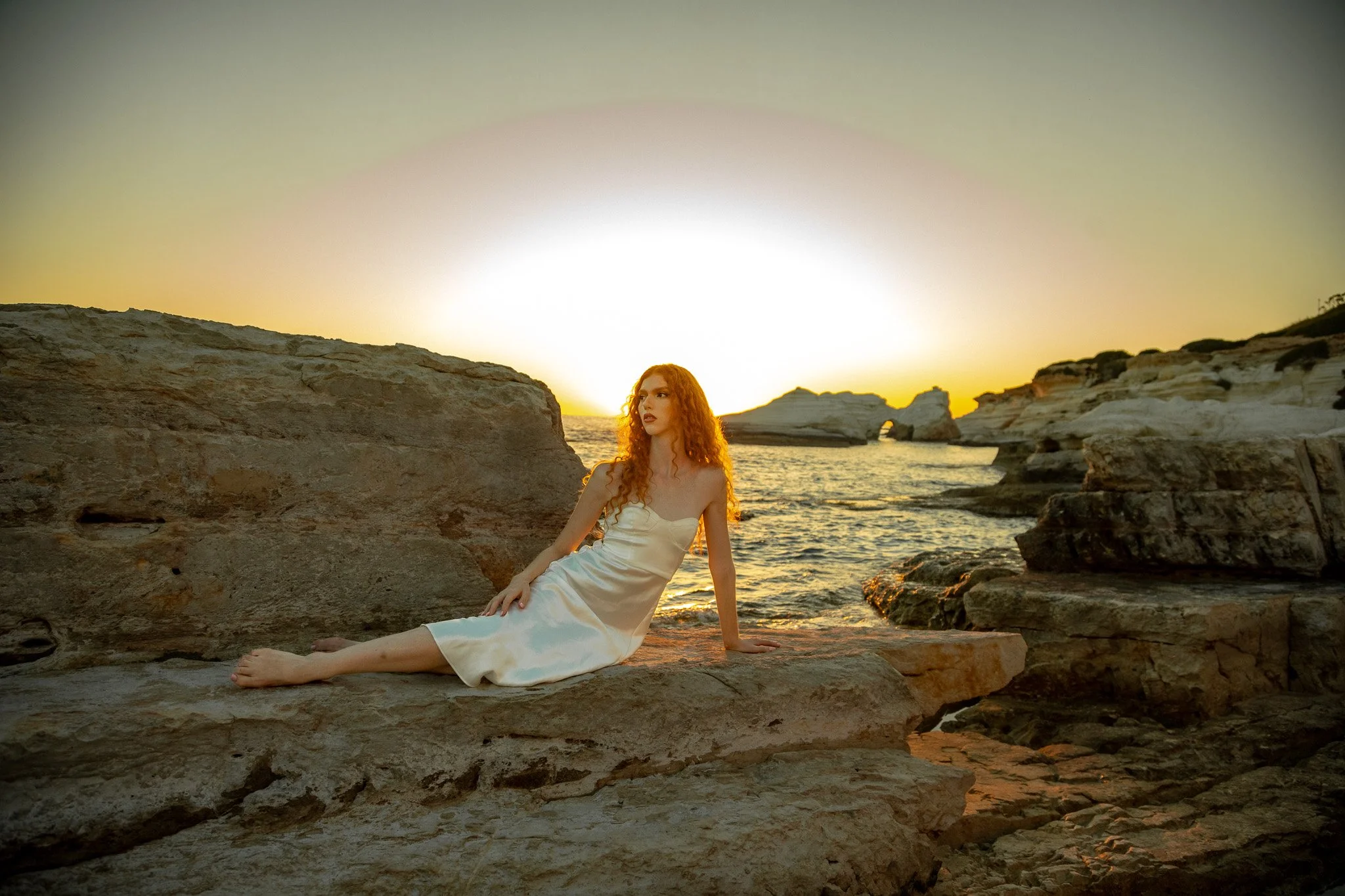 A woman with long curly red hair wearing a white dress sitting on rocks by the sea at sunset.
