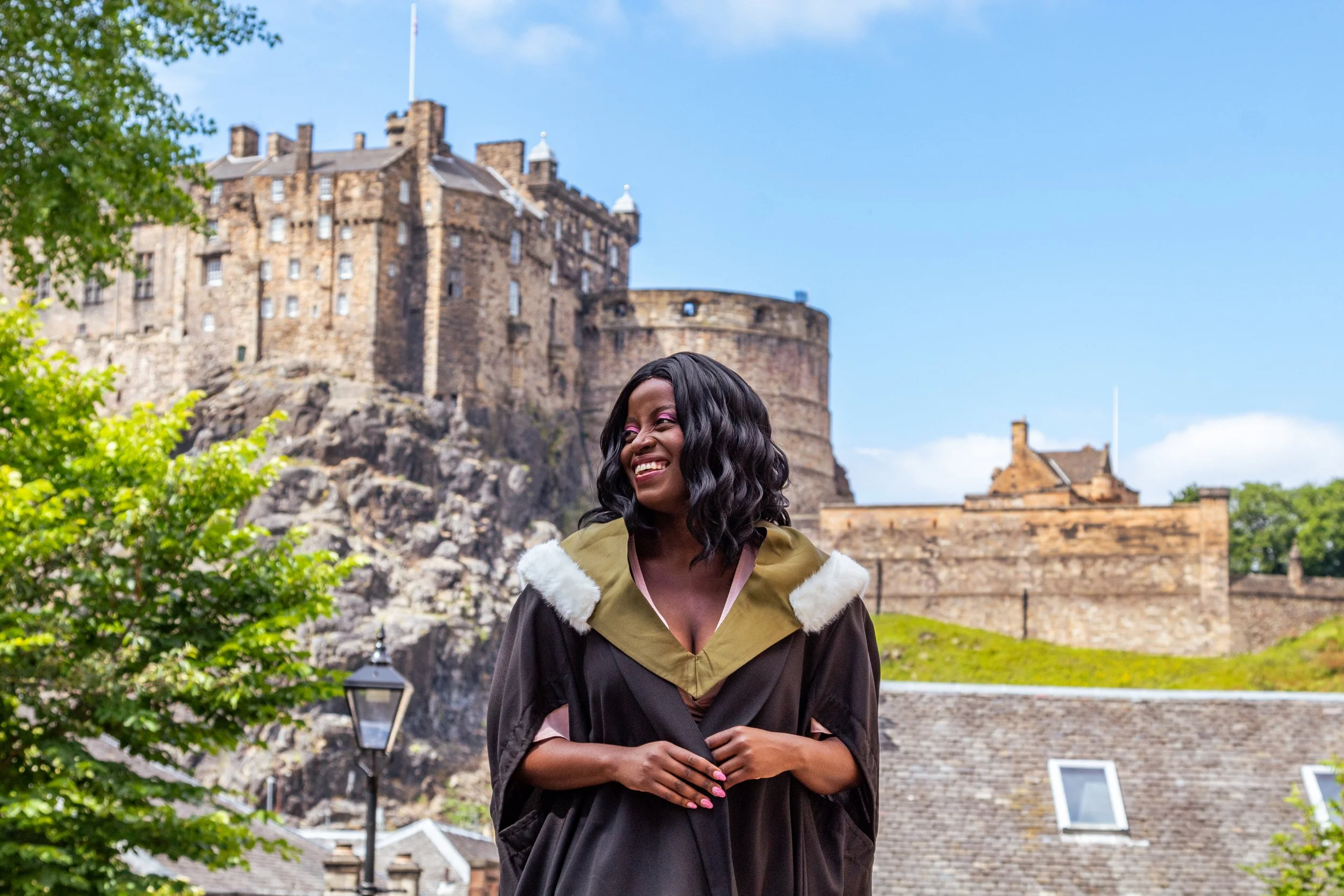 A woman in her graduation gown and cap stands outdoors with a castle in the background, smiling.