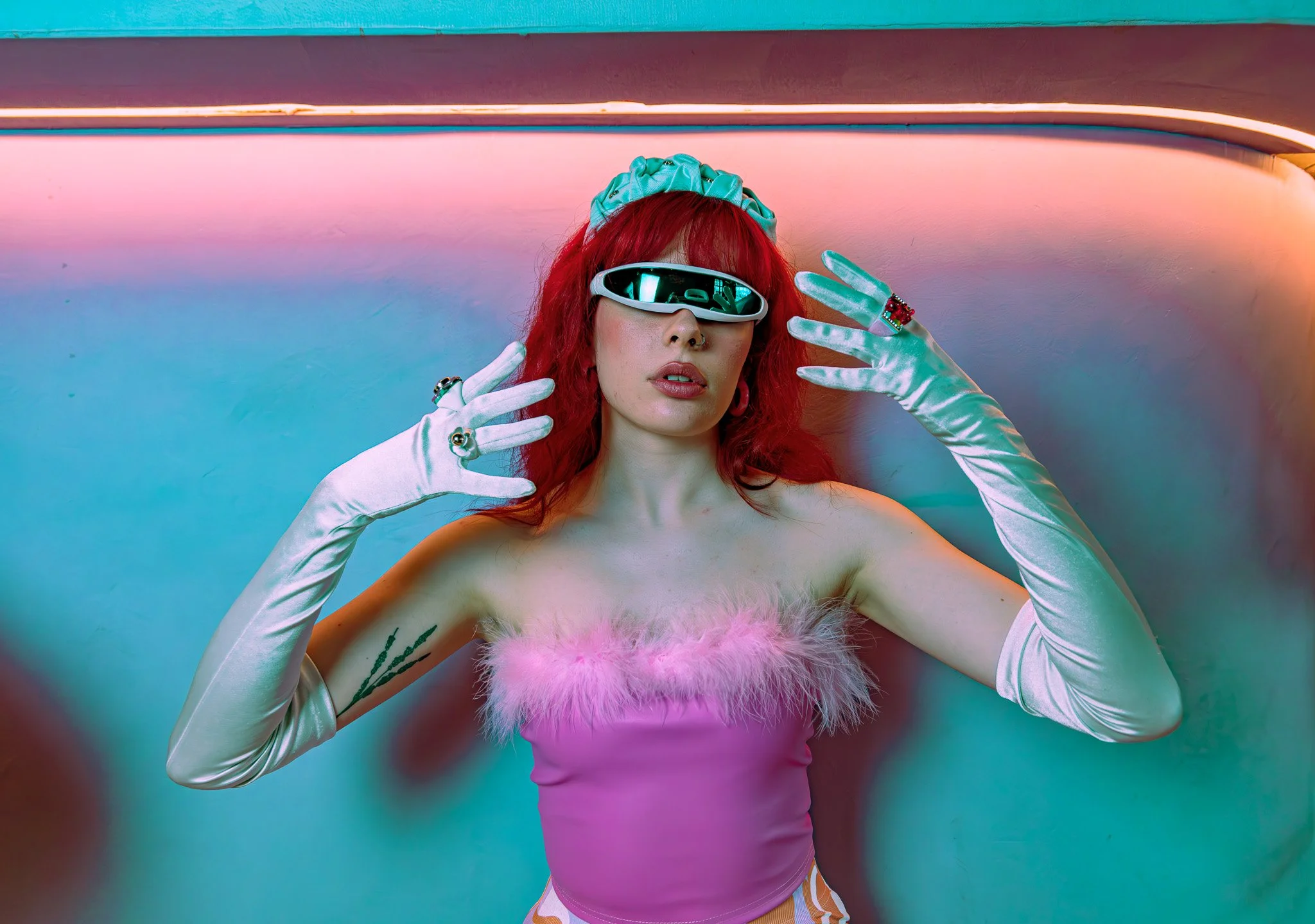 Woman with red hair wearing futuristic sunglasses, a pink top with feather details, white gloves, and jewelry, posing against a colorful, neon-lit background.