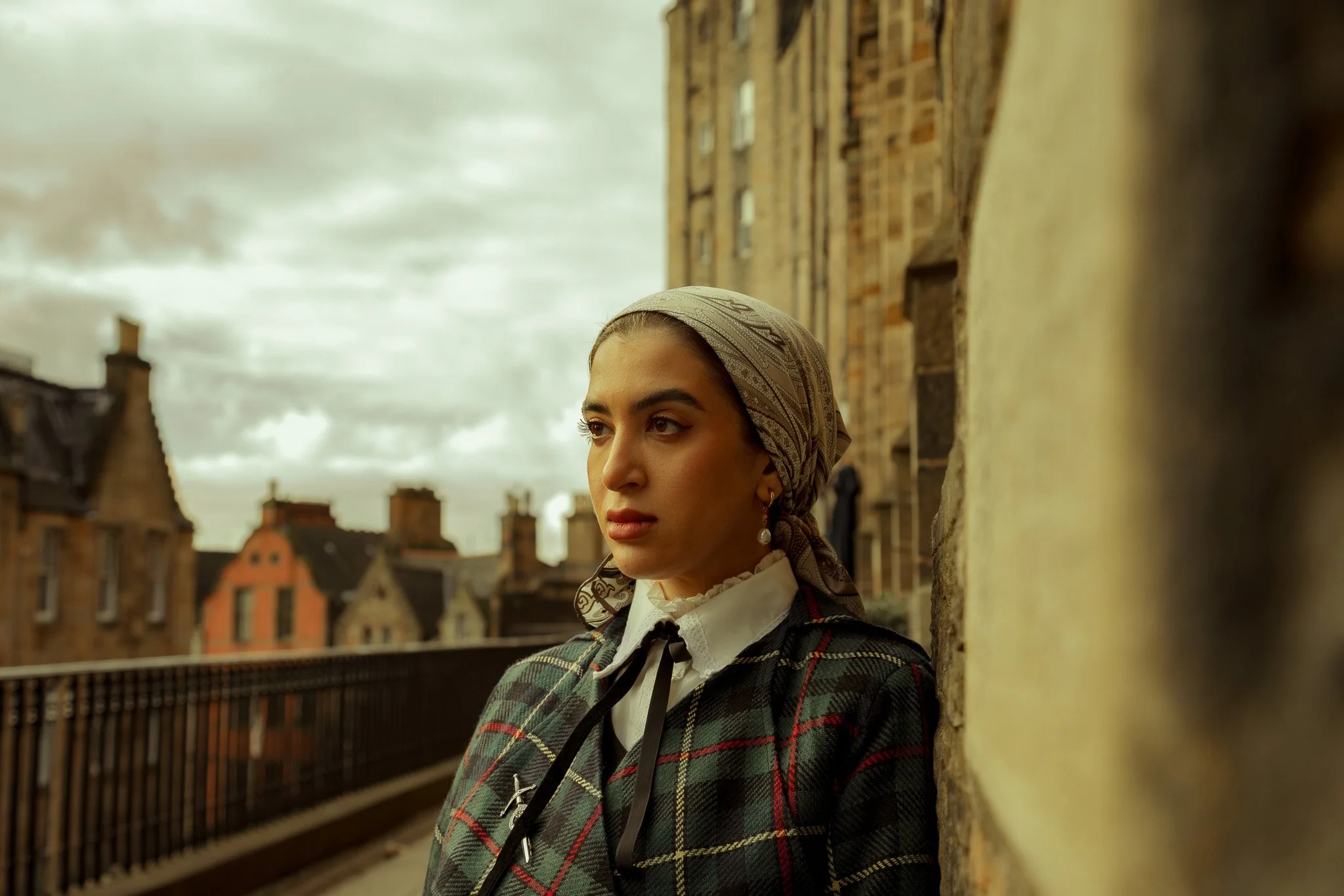 A young woman with a headscarf standing on a balcony overlooking historic brick buildings under a cloudy sky.