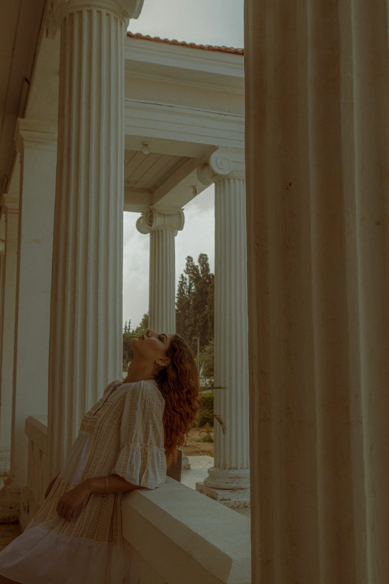 A woman with curly hair in a flowing dress leaning against a white balustrade outside a classical building with tall white columns.