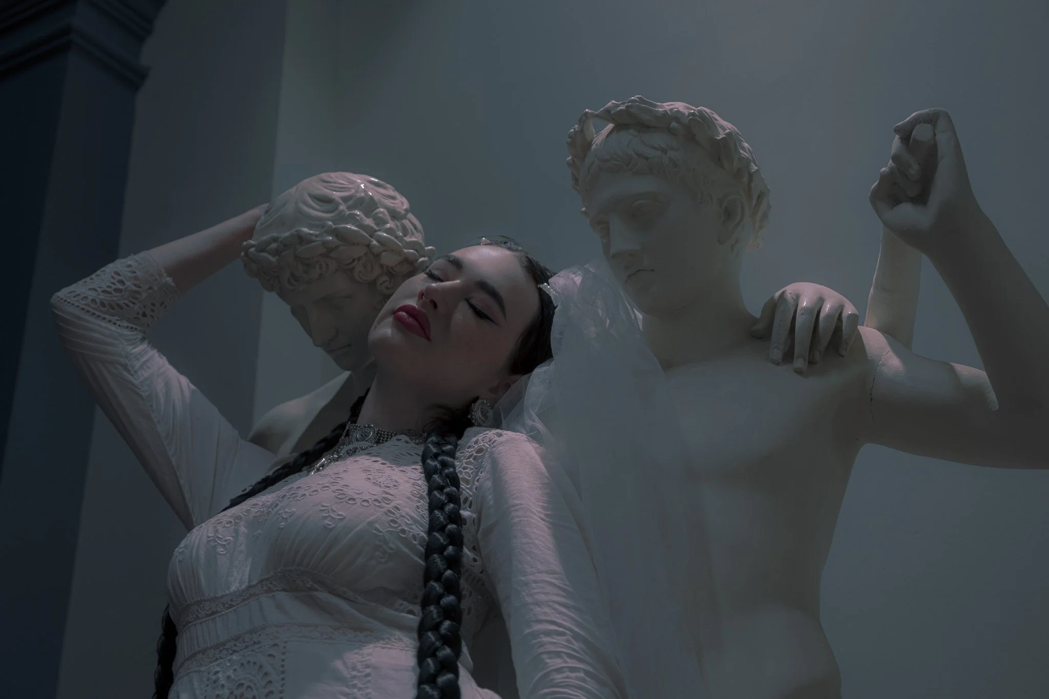 A woman with braided hair and bright red lipstick is resting her head against a classical statue of a man, surrounded by two other statues. The statues are white and appear to be made of marble, depicting figures from ancient Greek or Roman sculptures.