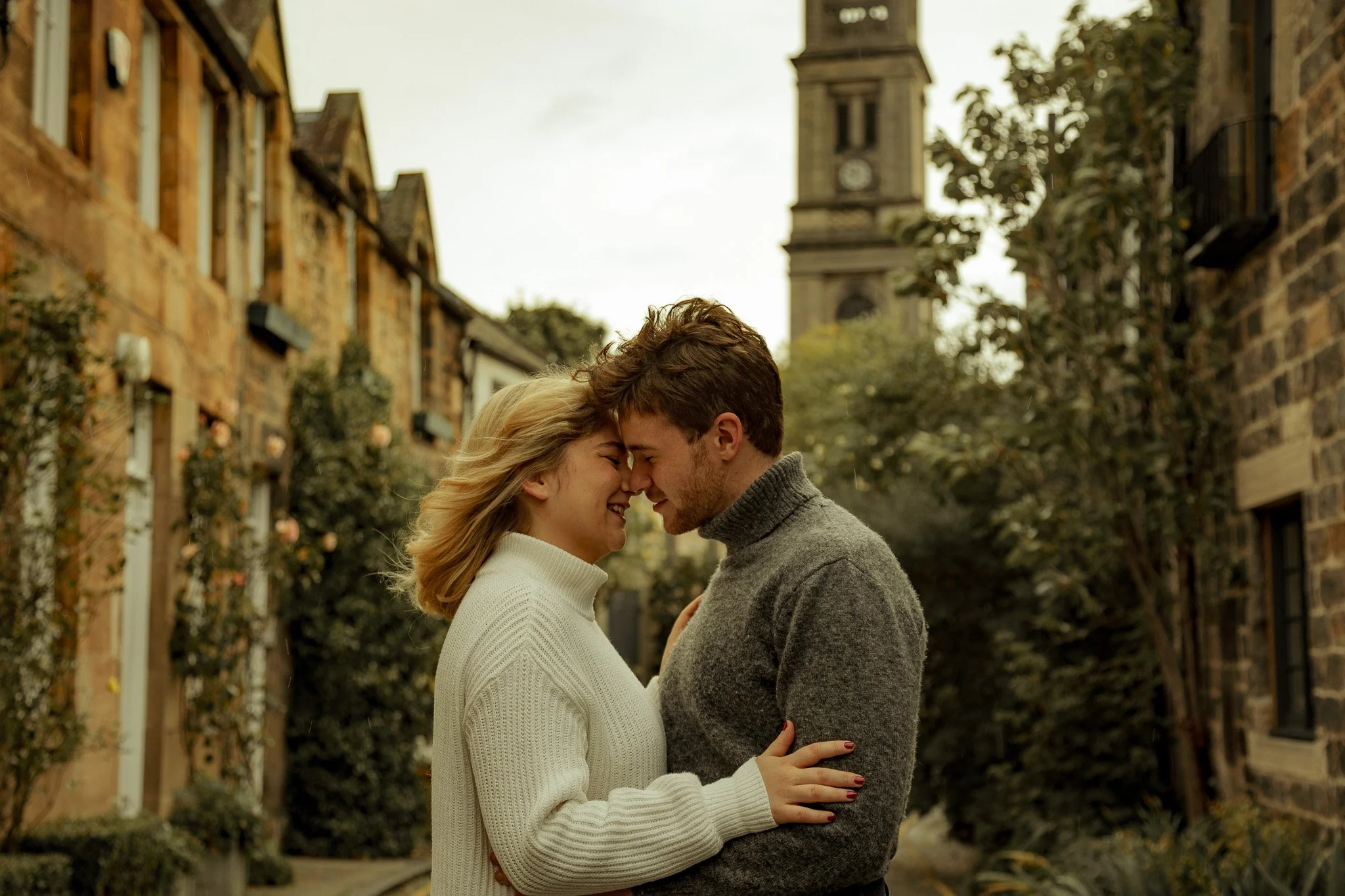 A young couple with their foreheads touching, smiling, in a narrow street with old brick buildings and a clock tower in the background, during daytime.