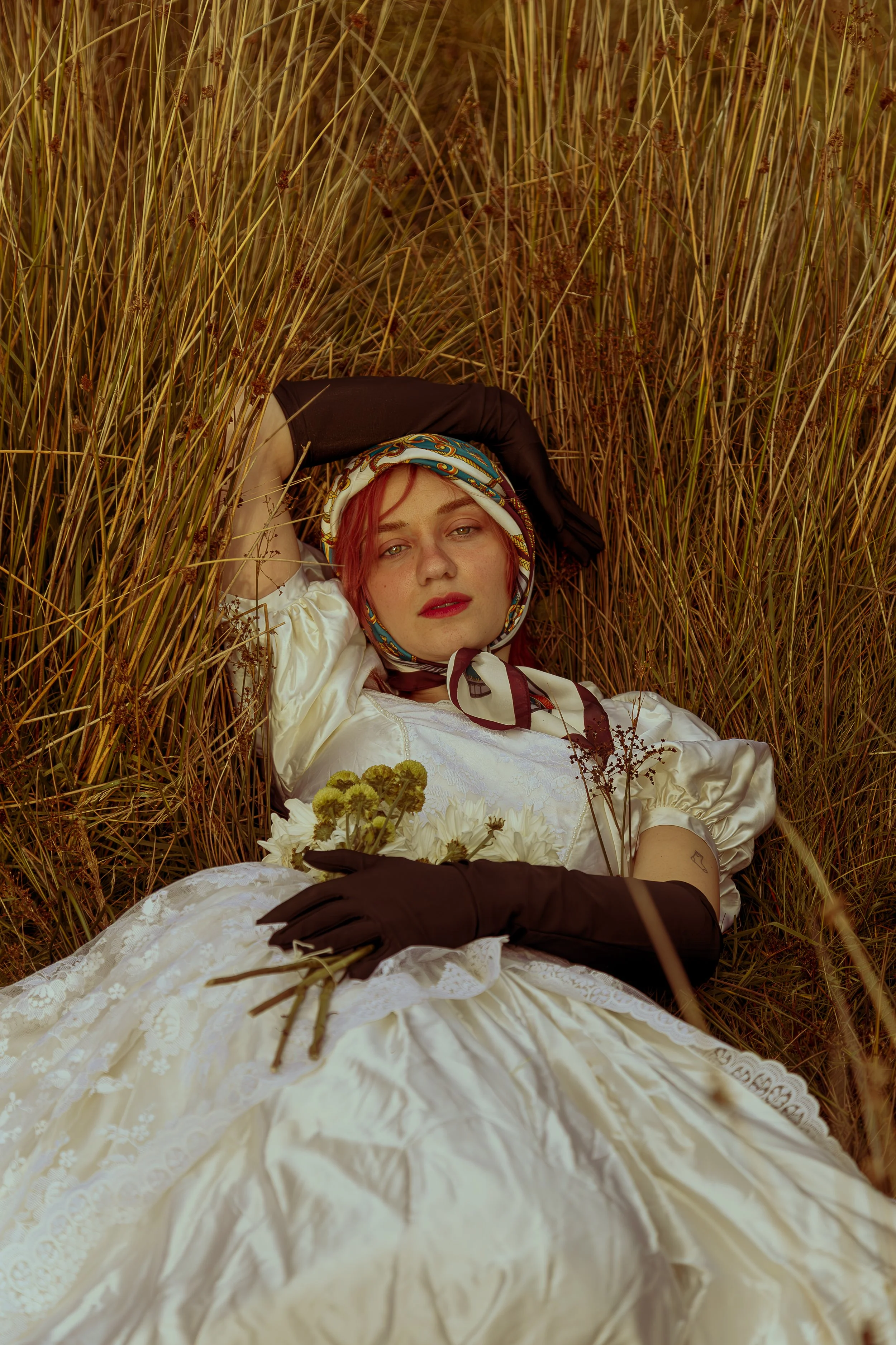 A young woman with red hair lying on her back in a field of tall grass, wearing a white vintage dress, black gloves, and a colorful scarf around her head, holding a small bouquet of flowers.