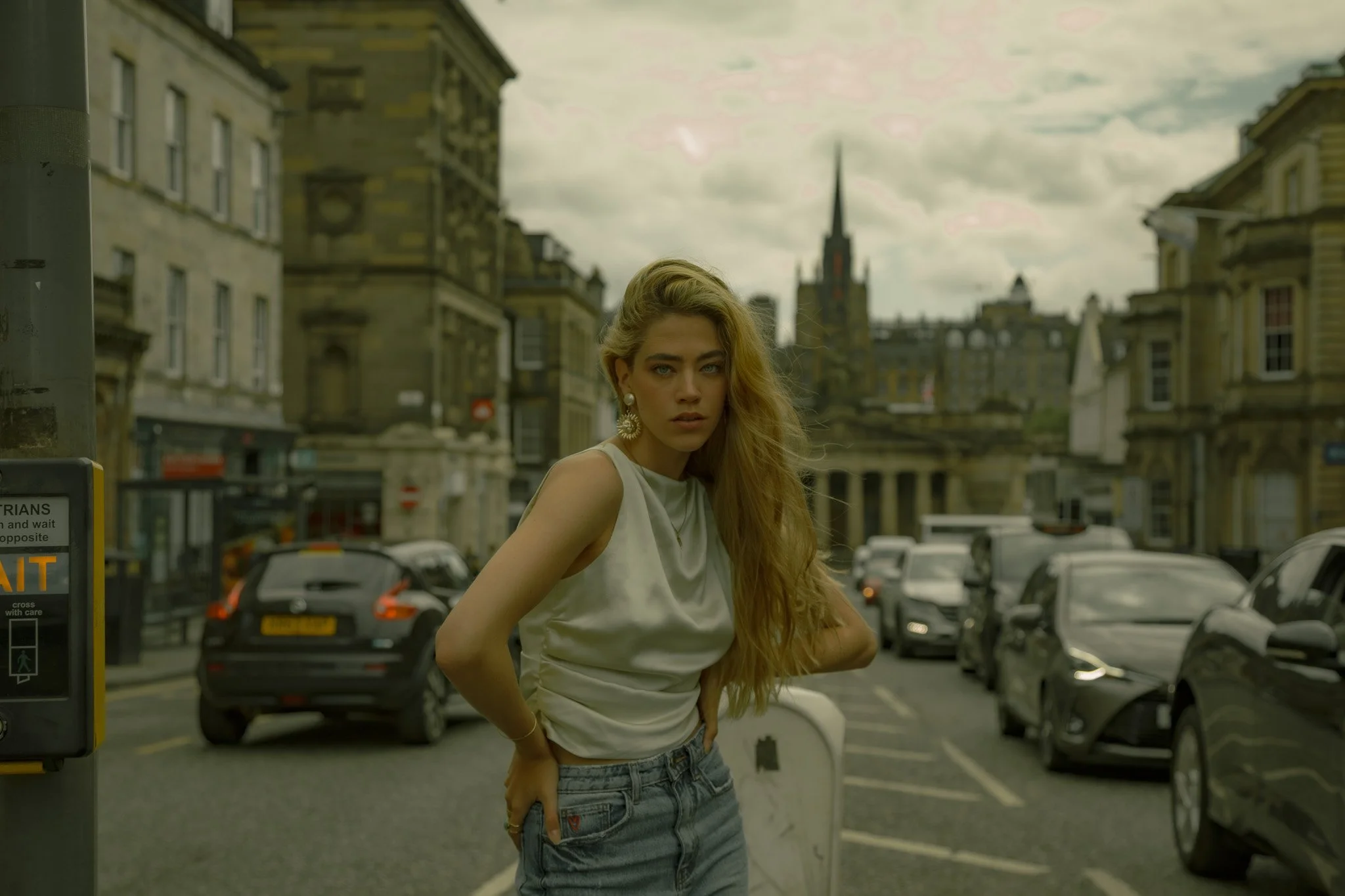 A young woman with long blonde hair standing on a city street with parked cars and historic buildings in the background, looking at the camera