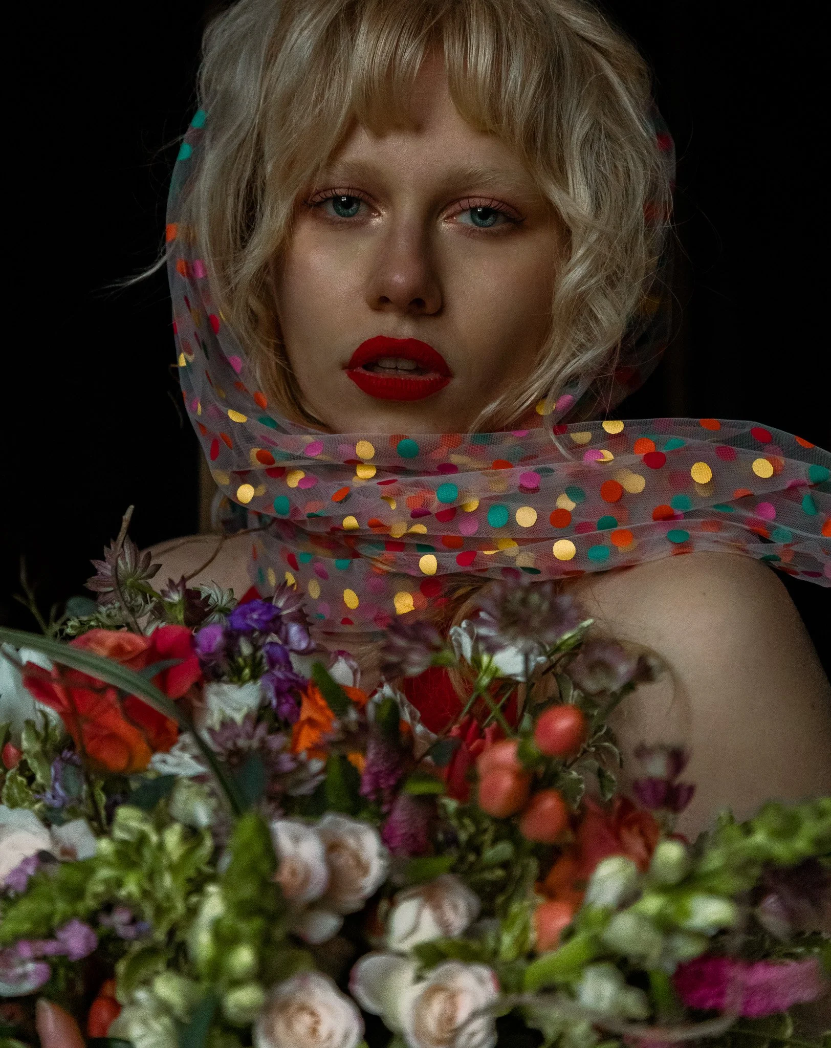 A blonde woman with wavy hair, wearing red lipstick, draped with a colorful, polka-dotted scarf, is surrounded by a large bouquet of various flowers against a black background.