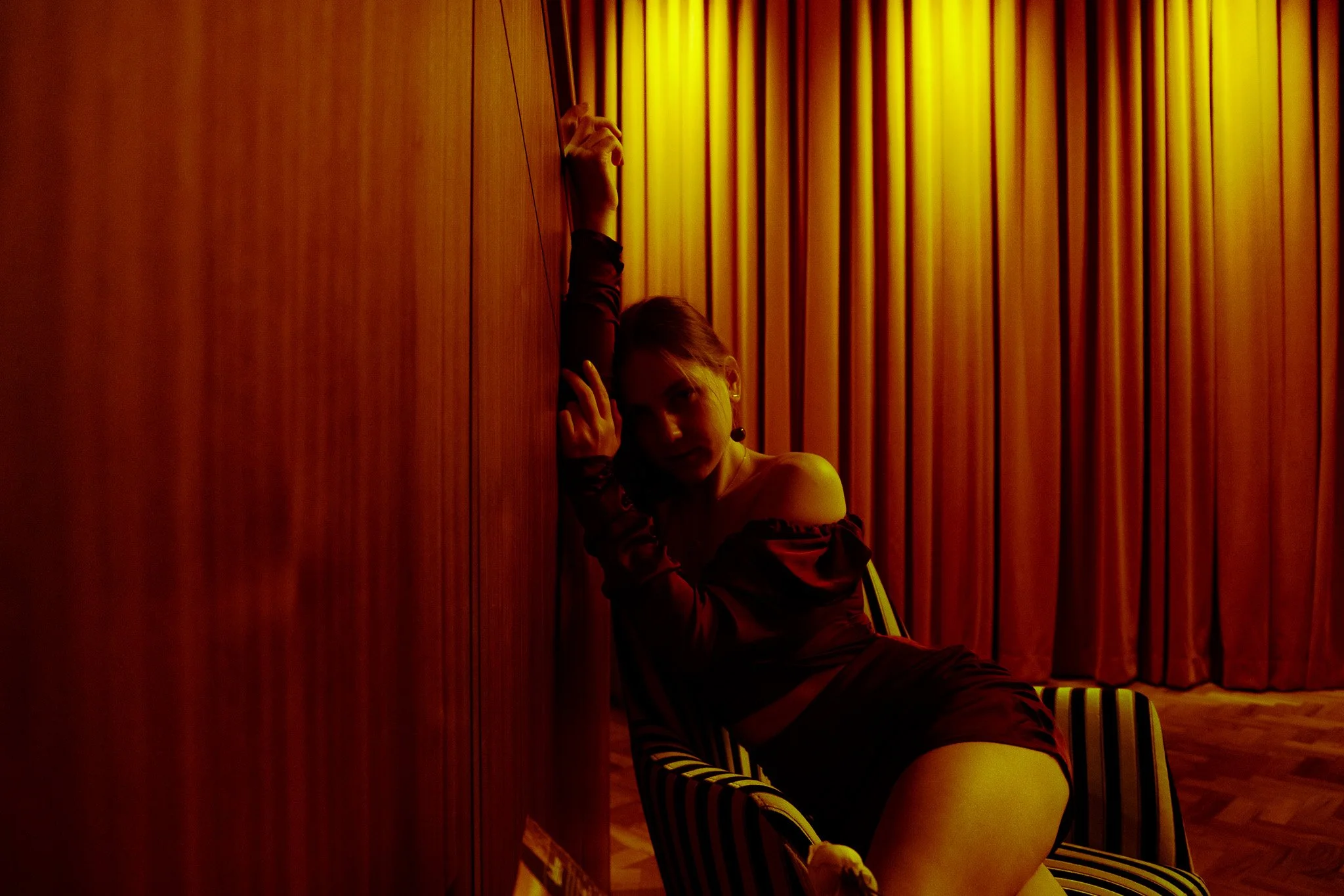 A woman in a dark, off-the-shoulder top leaning against a wooden wall, sitting on a striped chair in a dimly lit room with wooden paneling and curtains.