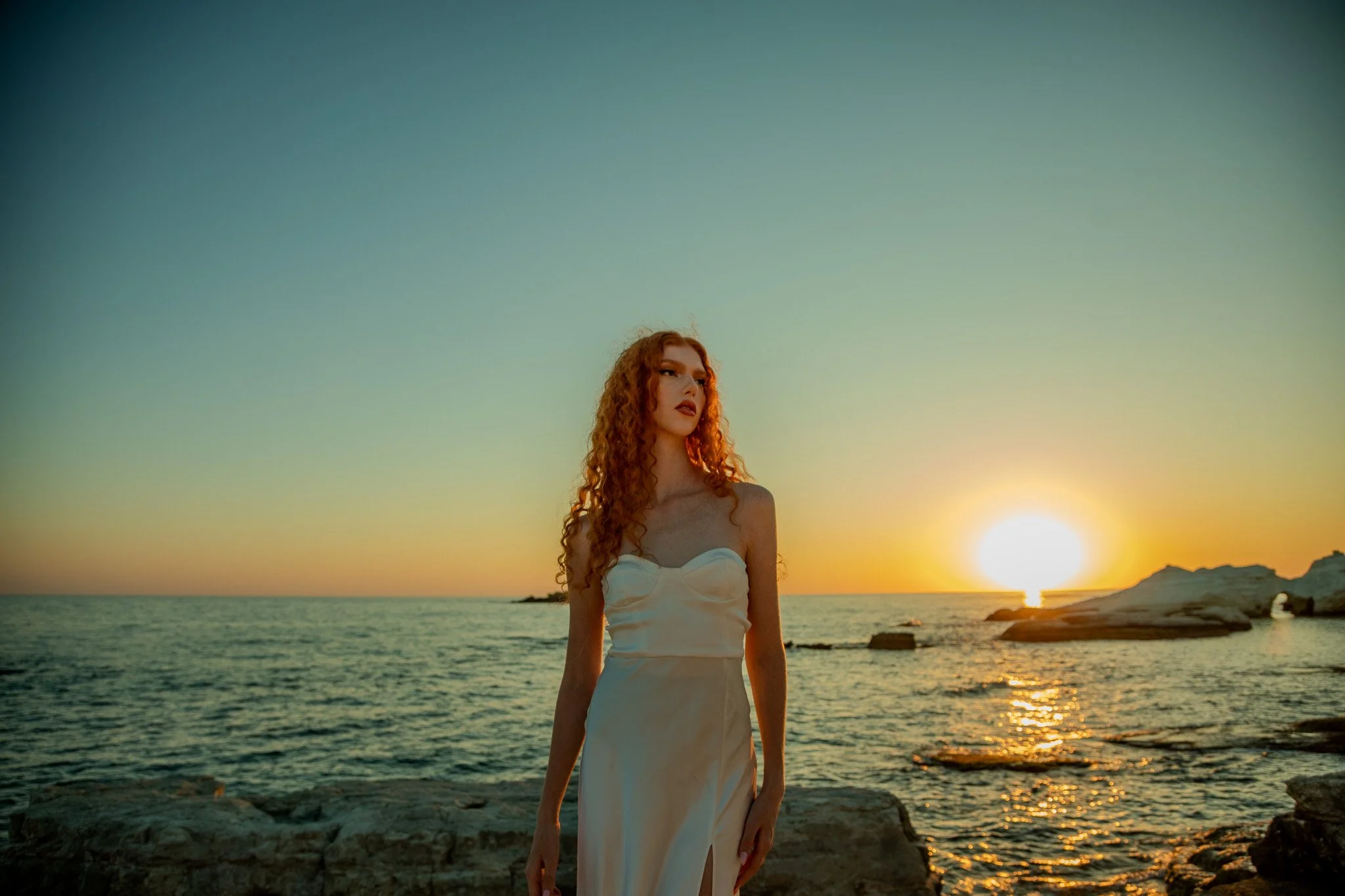 A woman with curly red hair in a white dress standing on rocks by the ocean at sunset.