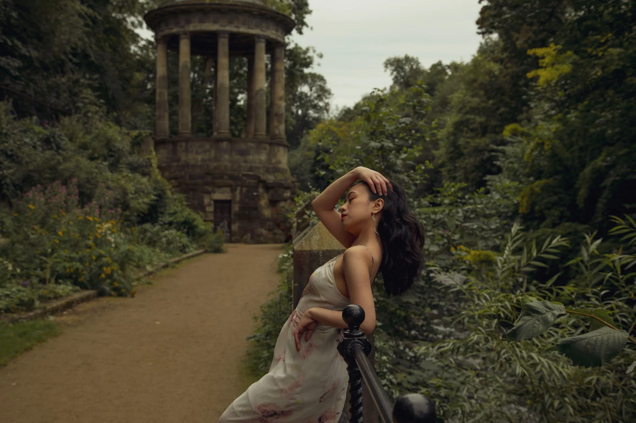 A woman in a cream-colored dress with floral patterns leans on a black railing with her eyes closed, holding her head with one hand, near a lush garden with a historic stone tower in the background.