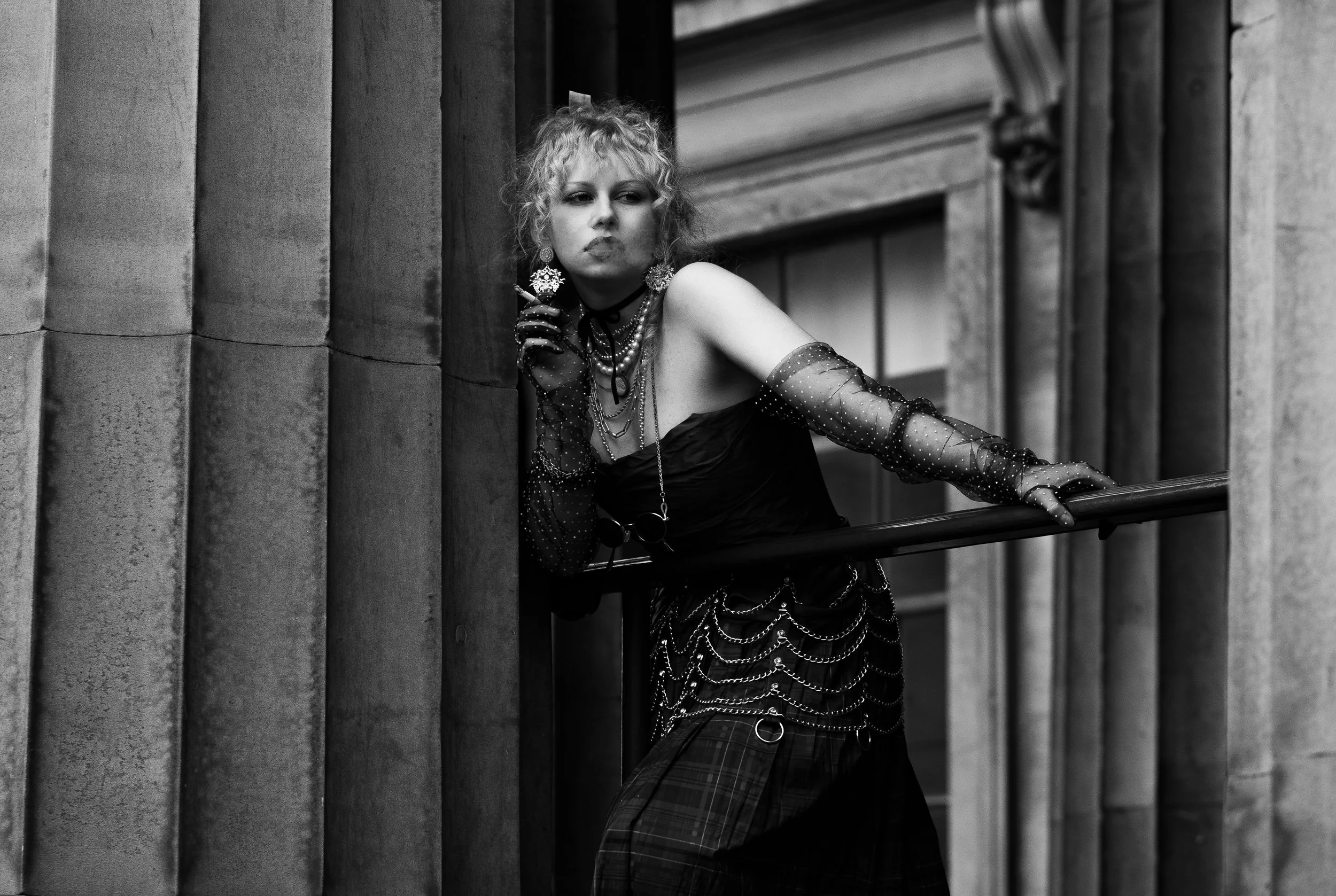 A woman with curly hair leaning on a railing outside a building, wearing a strapless dress, fishnet gloves, and layered jewelry in a black and white photo.