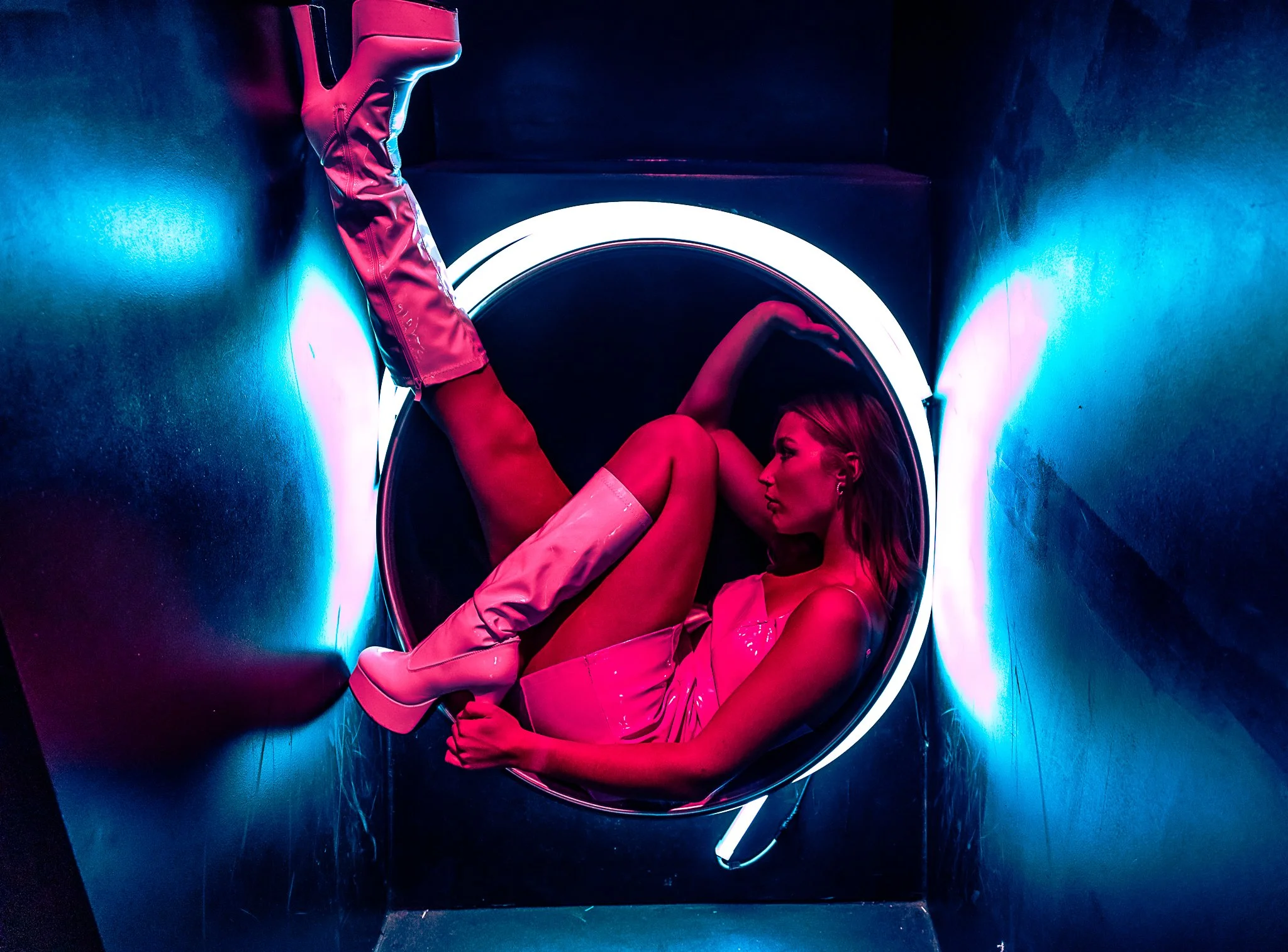 A woman in a pink dress and thigh-high boots lies inside a circular seating area with neon blue and pink lighting.