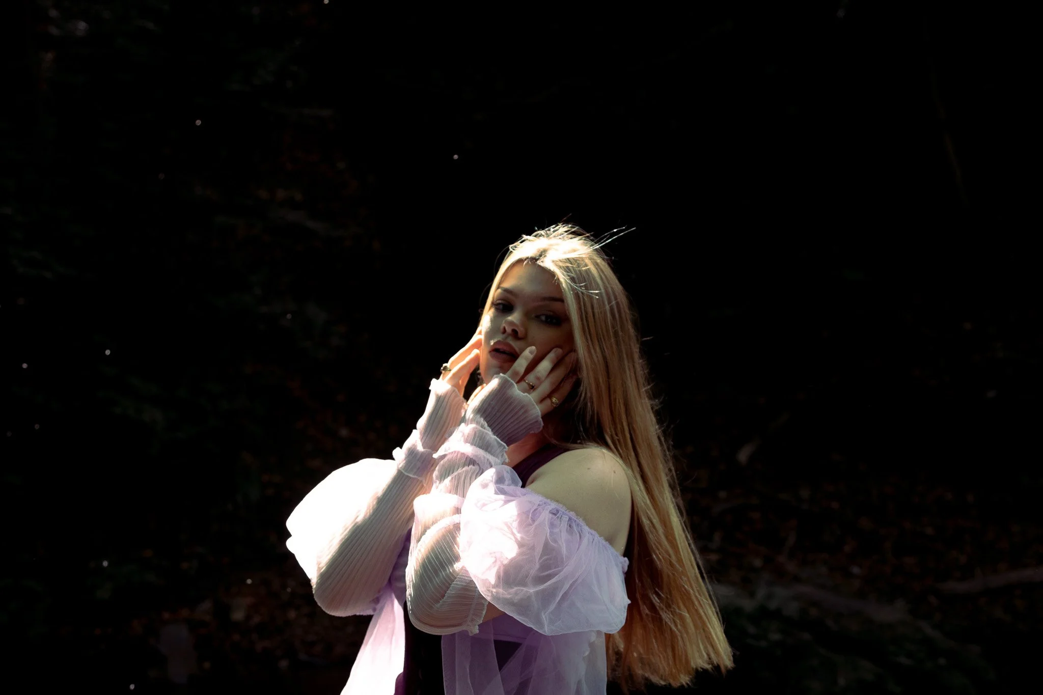 A woman with long blonde hair standing outdoors at sunset, wearing a black top and a sheer pink jacket with puffed sleeves, posing against a dark background.