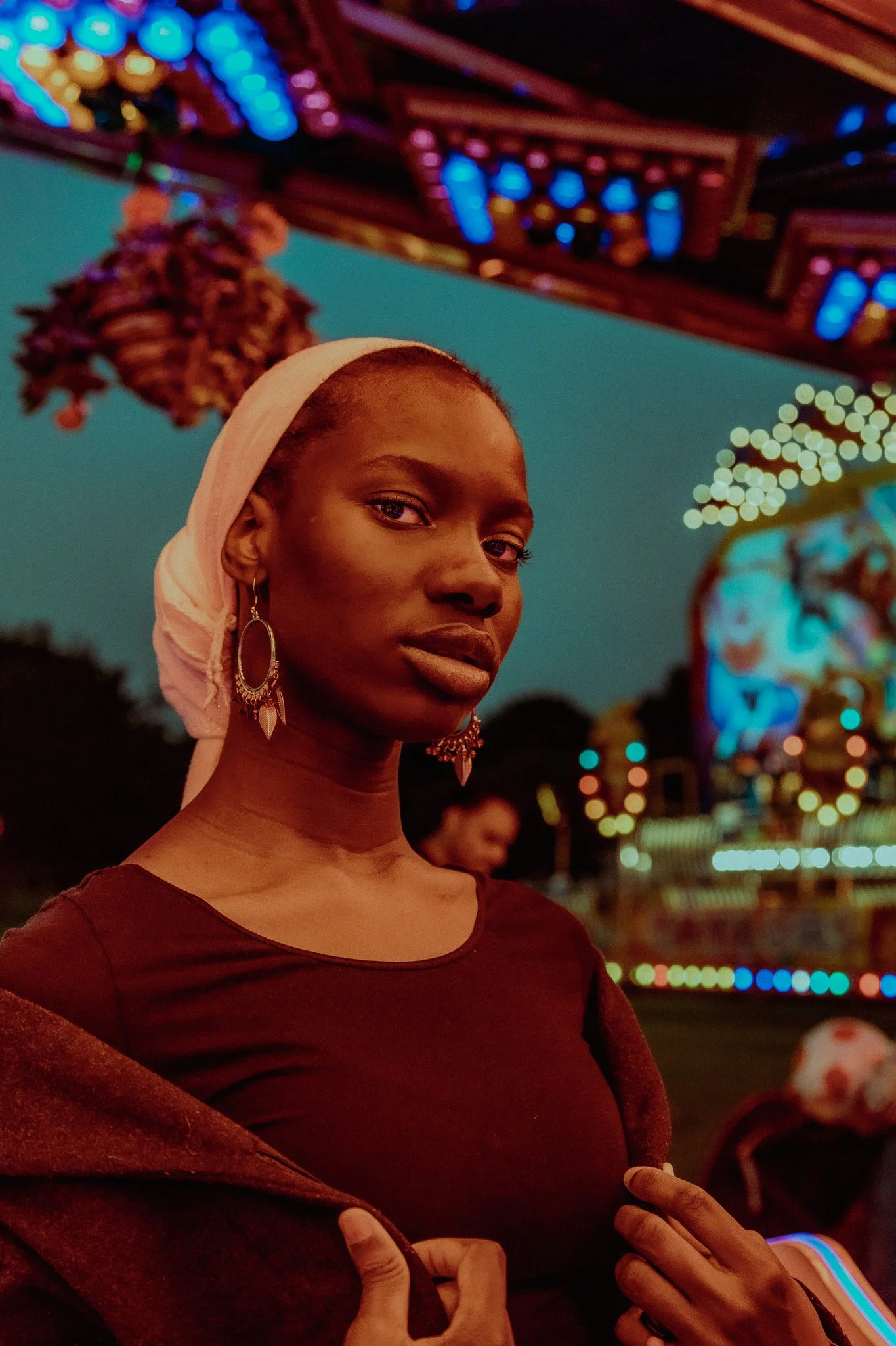 A woman with earrings and a white headwrap standing at a night fair with colorful lights and rides in the background.