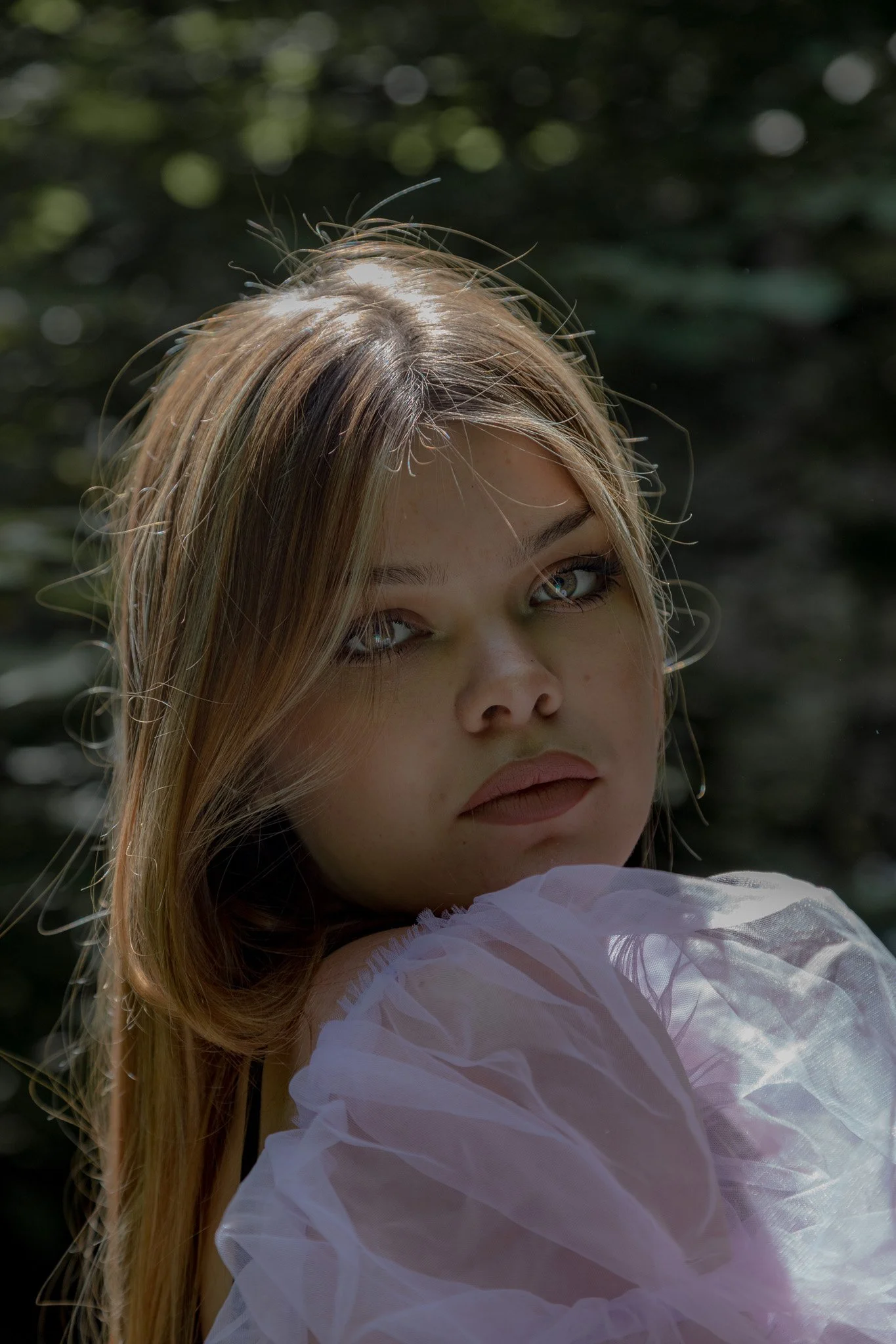 Close-up of a young woman with light brown hair, blue eyes, and fair skin, wearing a white, ruffled top, outdoors with blurred green foliage in the background.