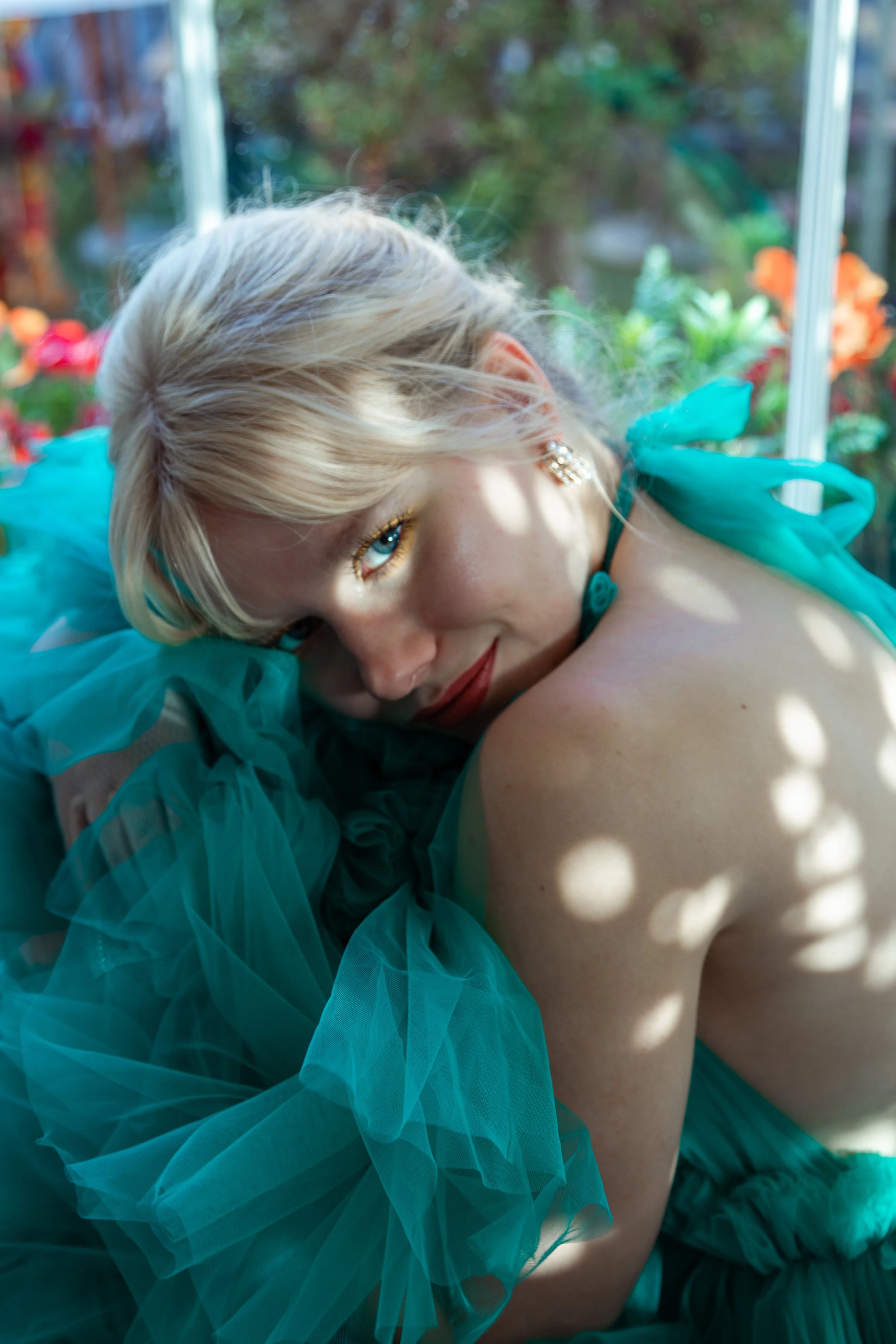 A woman with platinum blonde hair lying down in a garden, wearing a ruffled turquoise dress and earrings, with sunlight filtering through the leaves creating dappled light on her skin.