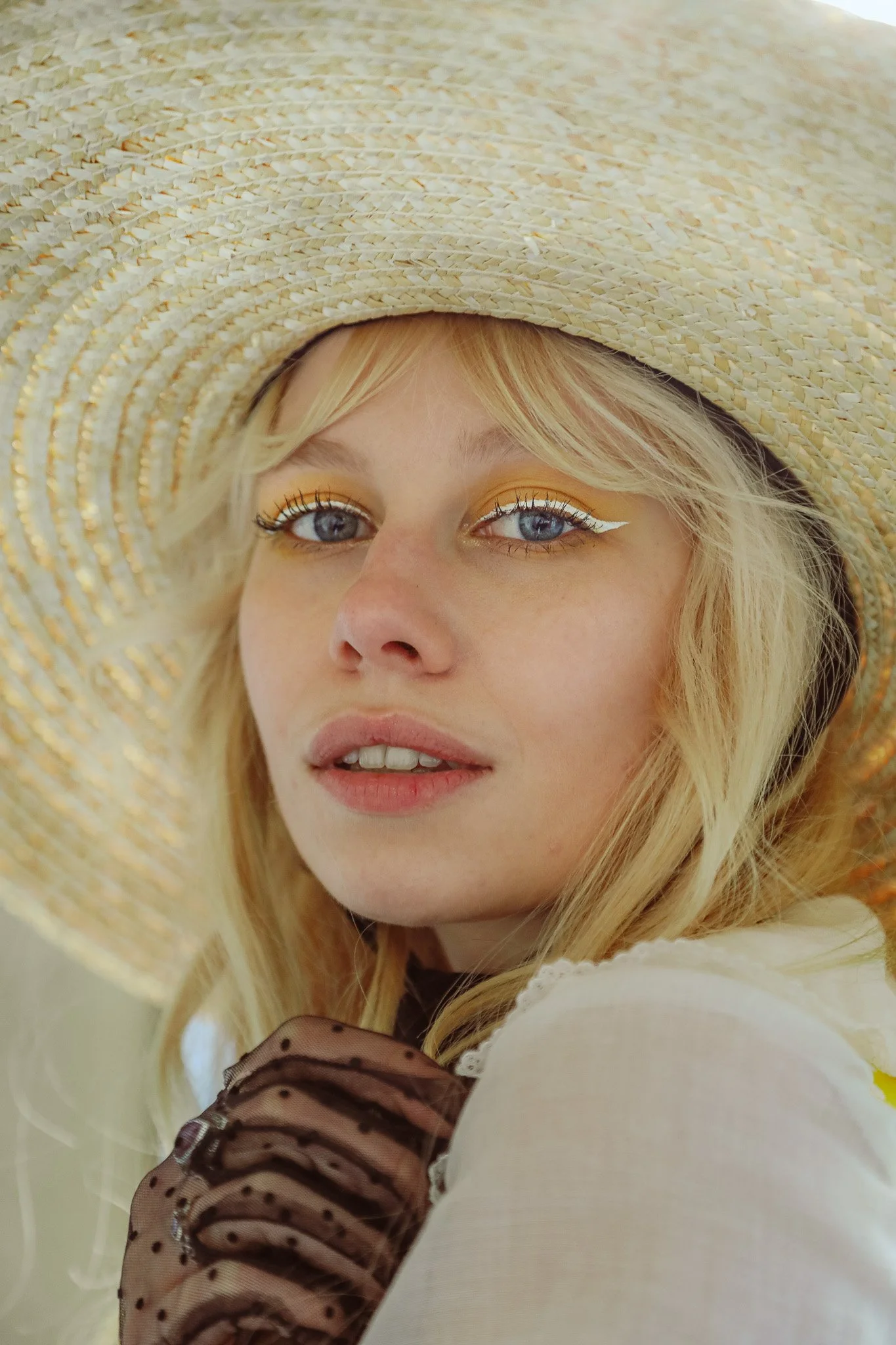 A woman with blonde hair wearing a large straw hat, sheer black polka dot gloves, and white clothing, looking at the camera with blue eyes and makeup with yellow eyeshadow and white eyeliner.