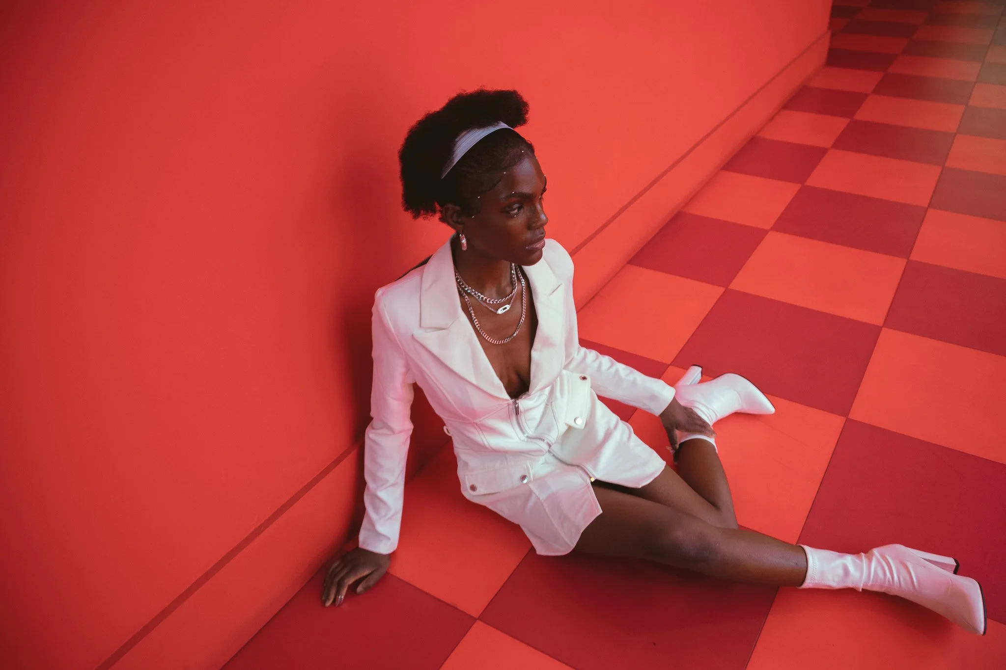 A woman in a white suit and boots sitting on a checkered red and pink floor, leaning against a red wall, with jewelry and a headband.