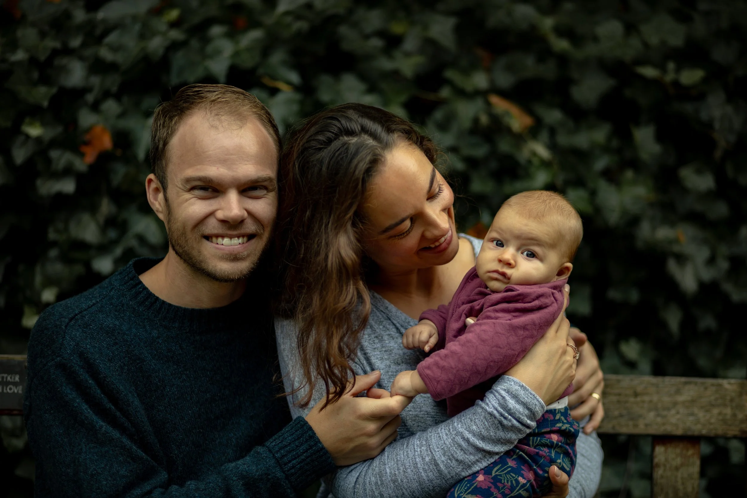 A happy family of three portrait outdoors, a man, woman, and baby, with a dark leafy background. The woman is holding the baby, and the man is smiling at the camera.