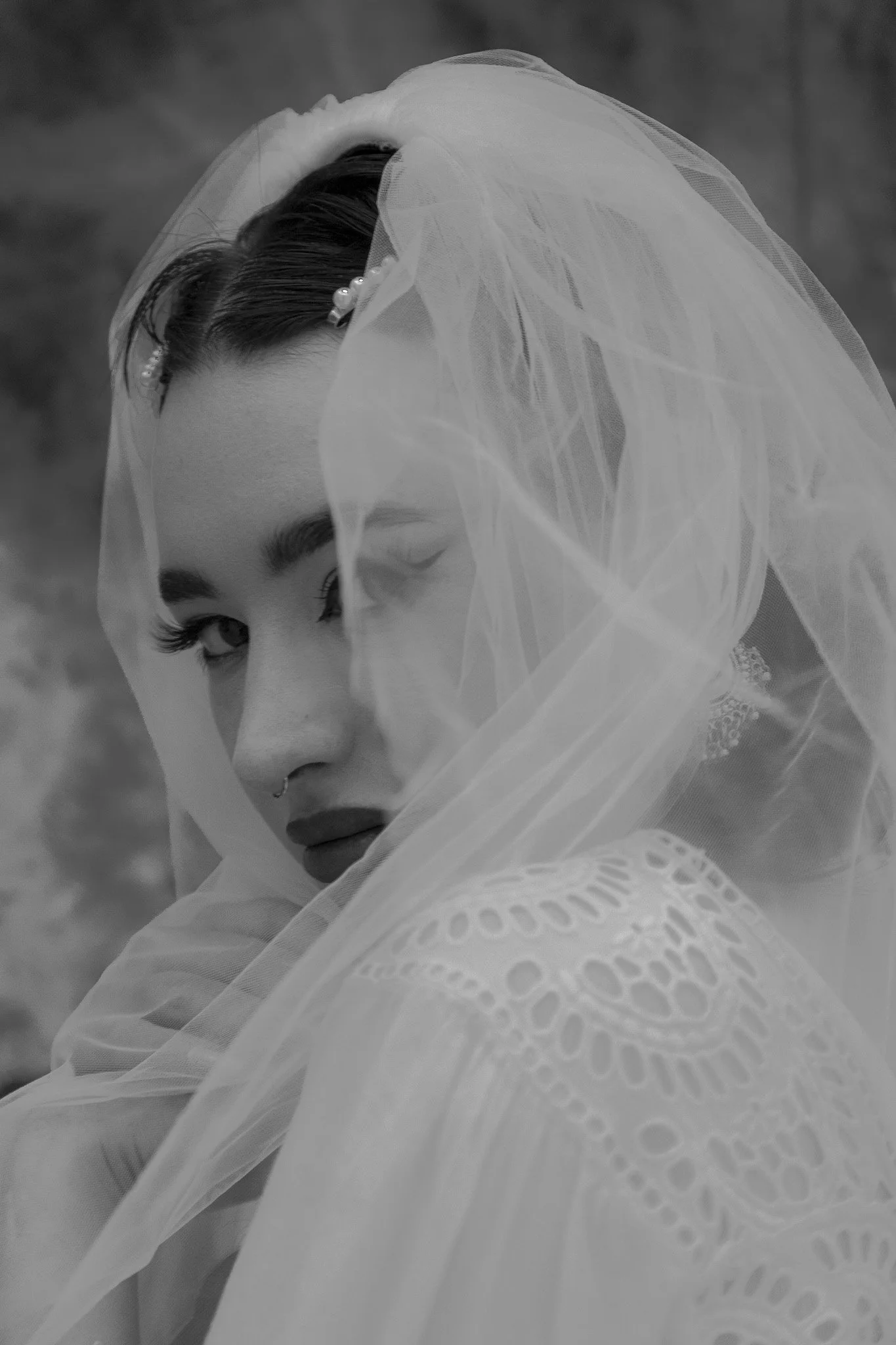 A woman with dark hair, makeup, and a septum piercing, partially covered with a sheer veil, posing with a confident expression.