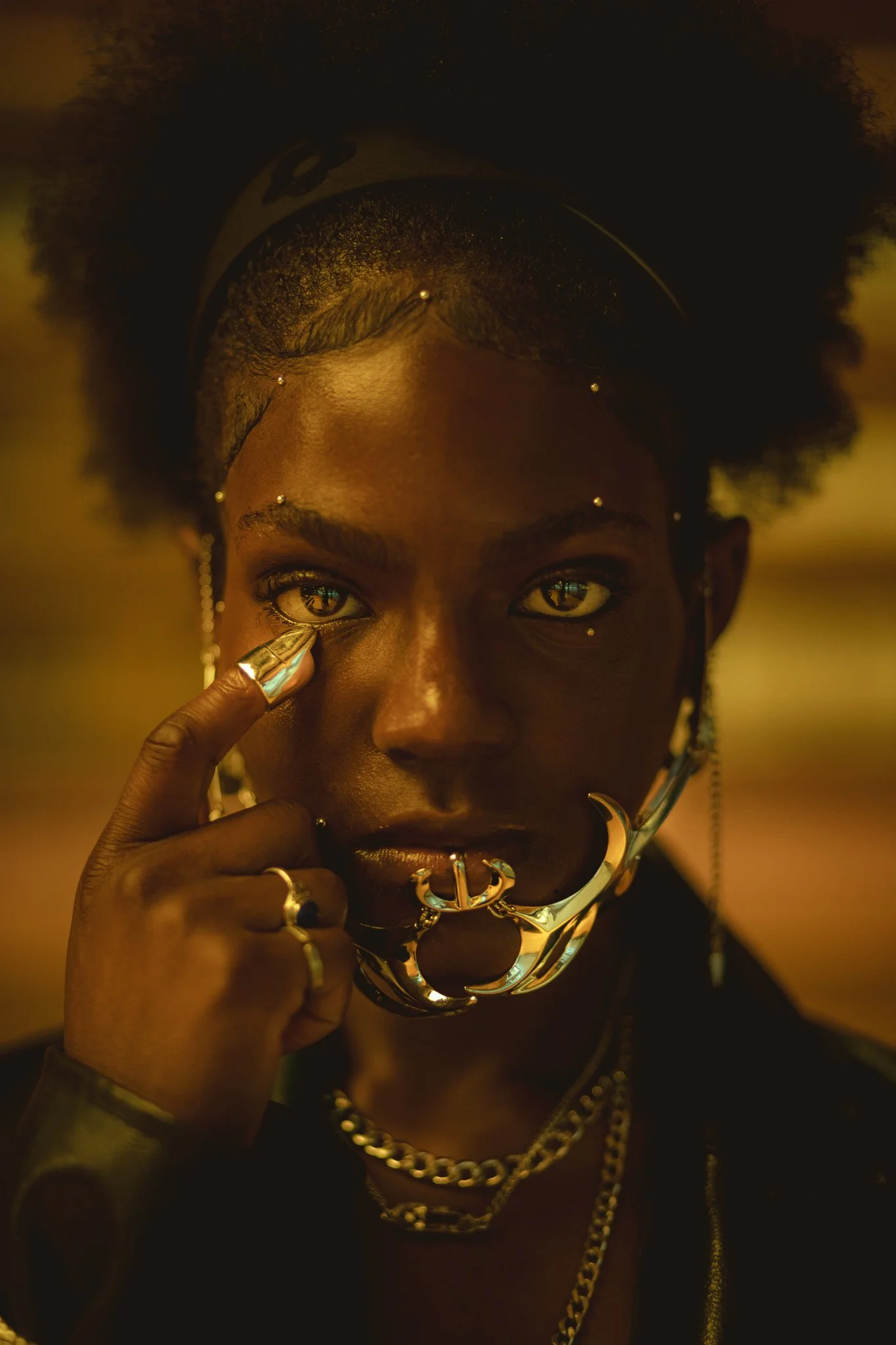 A woman with dark skin and fiery eyes, adorned with metallic jewelry including a large nose ring, multiple rings, earrings, and layered necklaces, touching her face with her finger, with a golden-hued background.