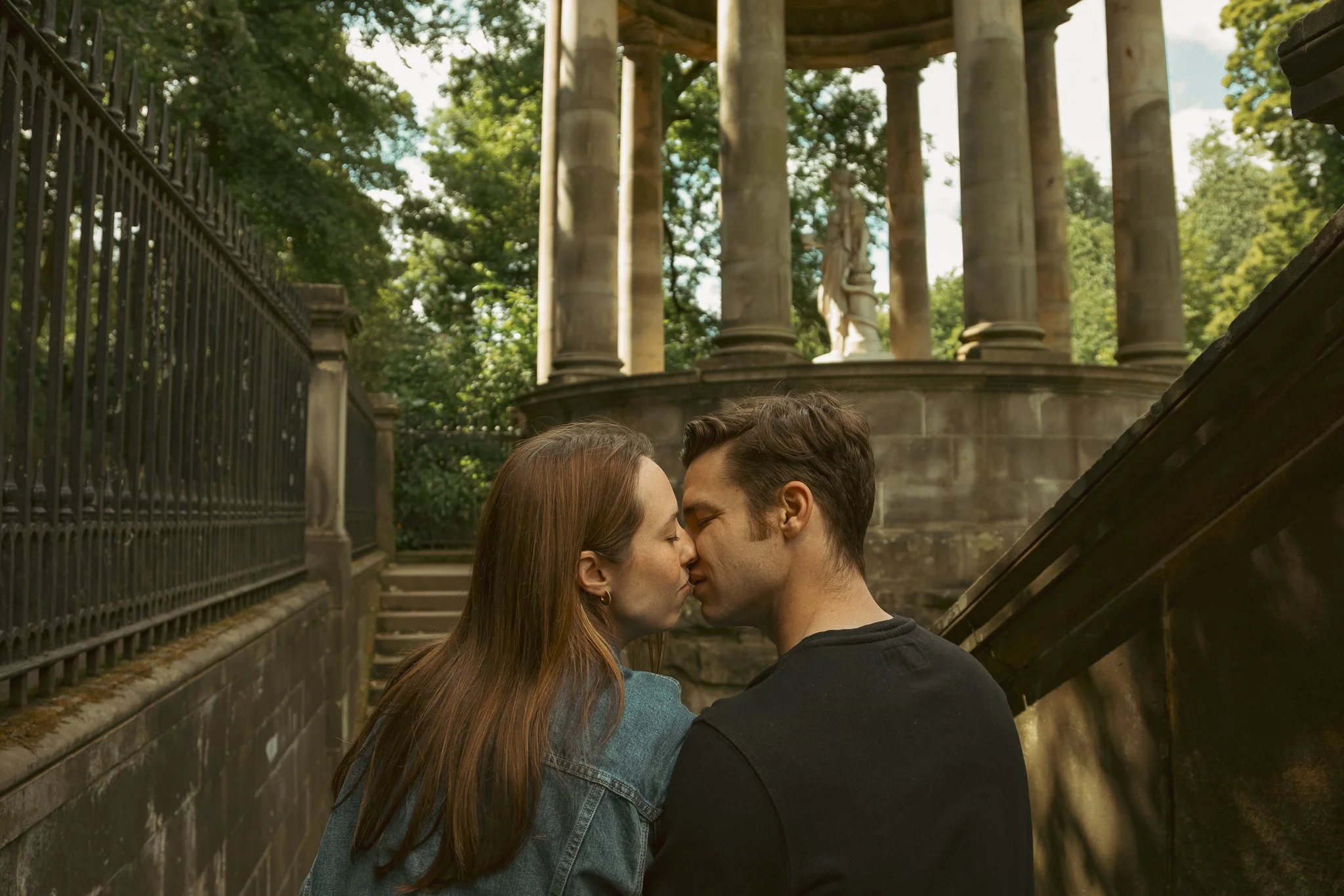 A couple kissing outdoors on a stone staircase, with an ornate stone structure and statues in the background surrounded by green trees.