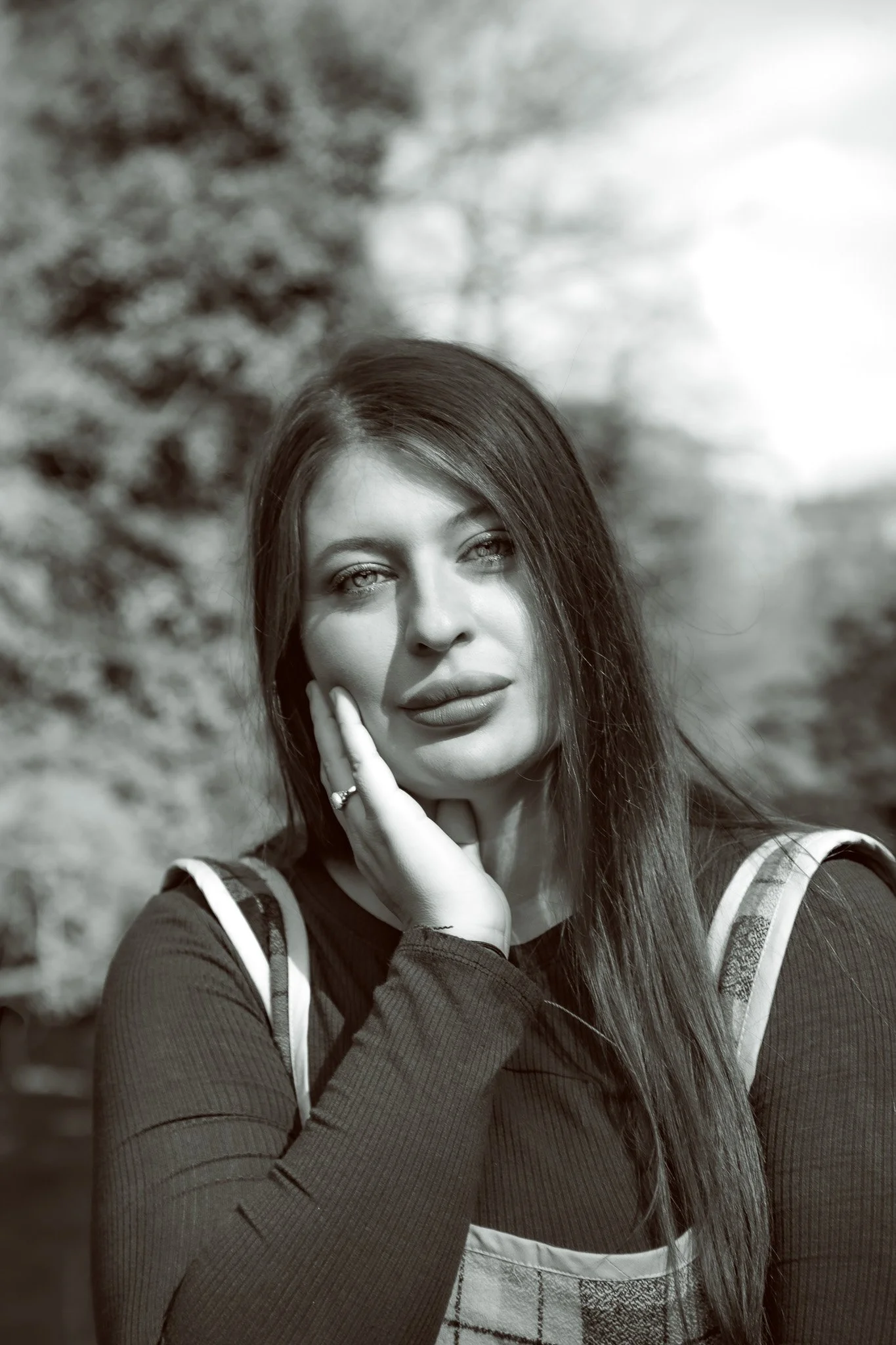 A black and white photograph of a woman with long hair, posing outdoors with her hand touching her face, with trees in the blurred background.